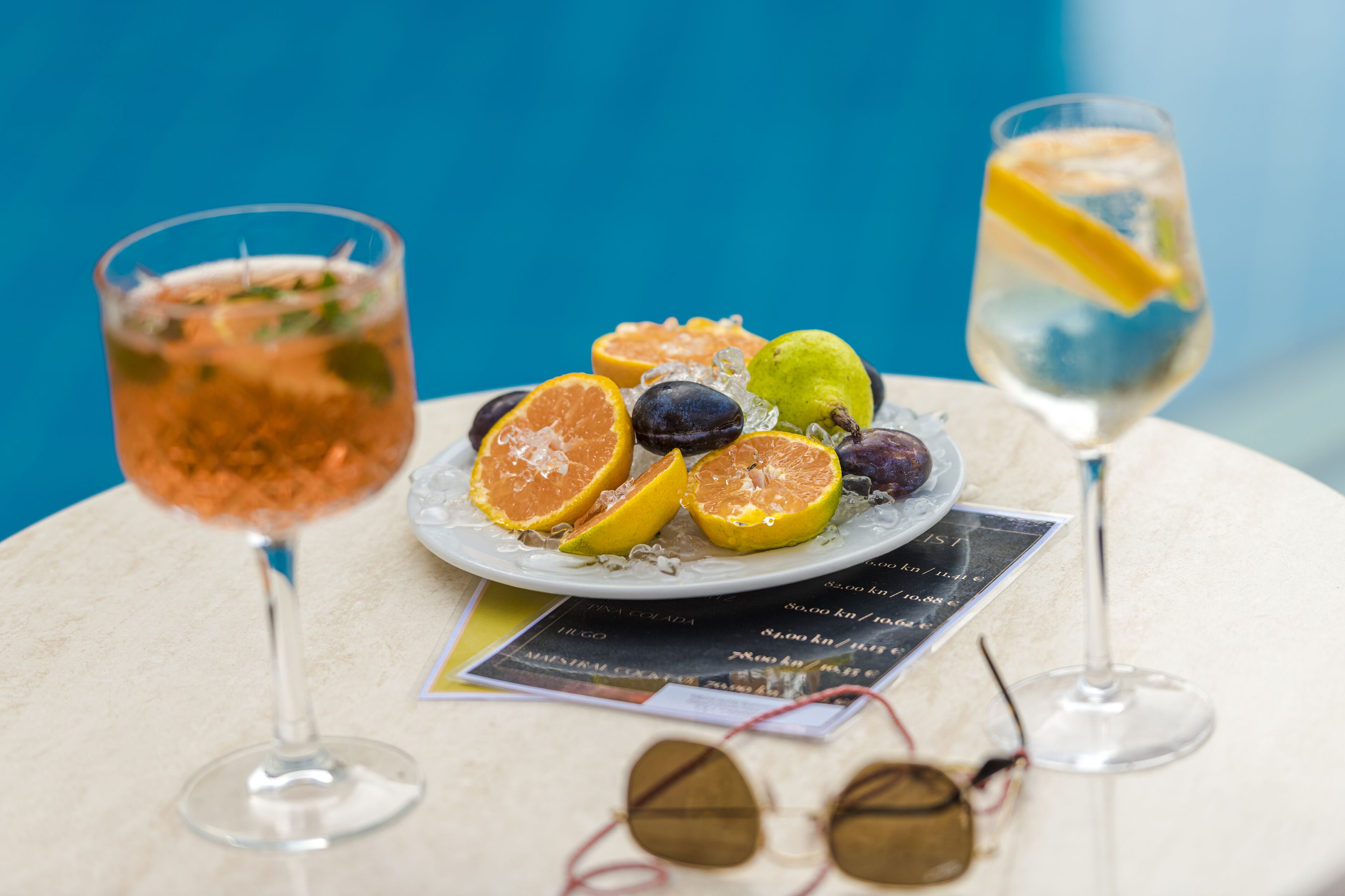 Two glasses of cocktails, a plate with fruit slices, and a menu on a table.