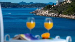 A table with two wine glasses and a plate of food, overlooking a coastal view with buildings and mountains in the background. - Hotel Komodor (Dubrovnik)