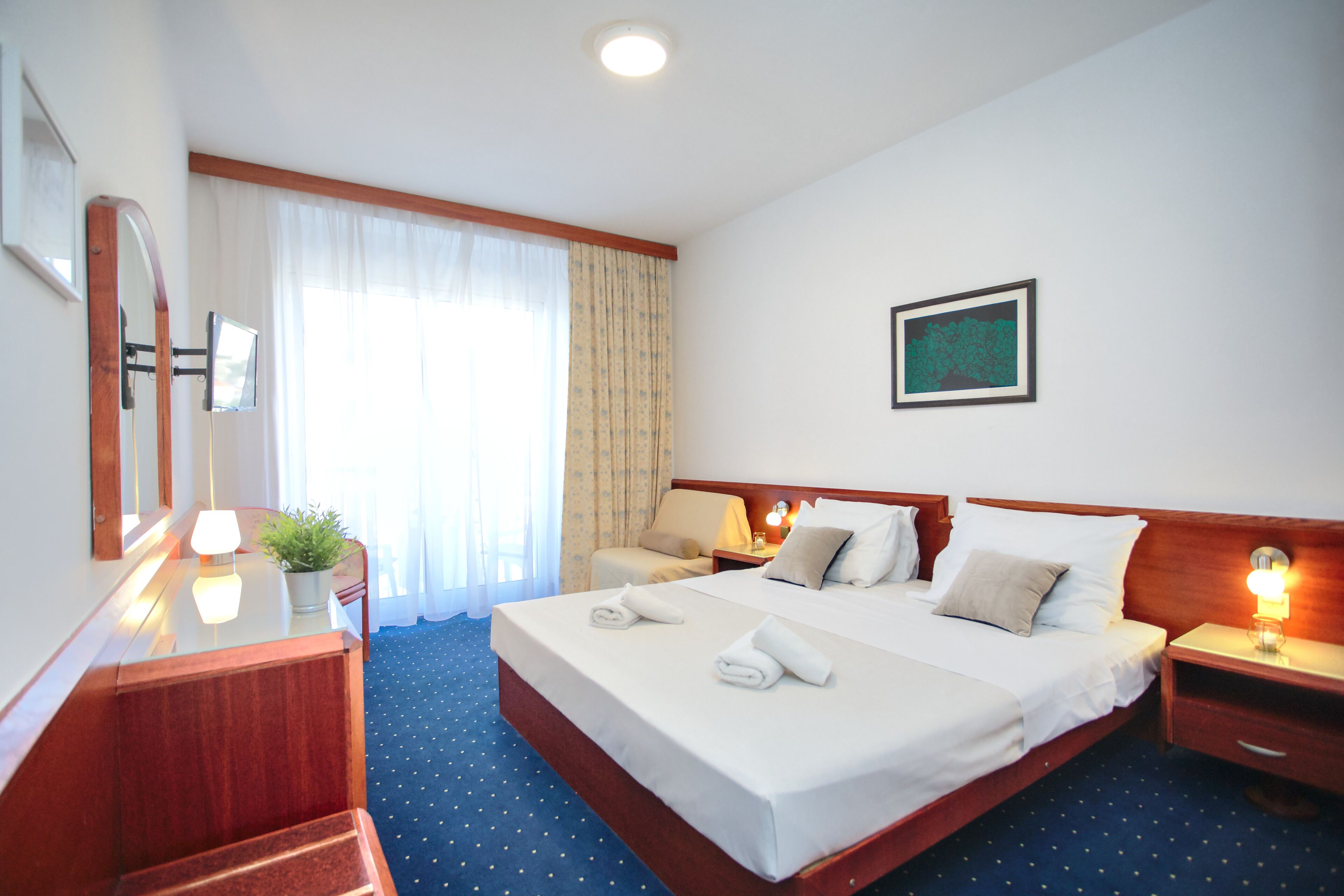 Une chambre d’hôtel avec un grand lit, deux tables de chevet avec des lampes, un bureau avec une chaise, une plante en pot et une fenêtre avec des rideaux.