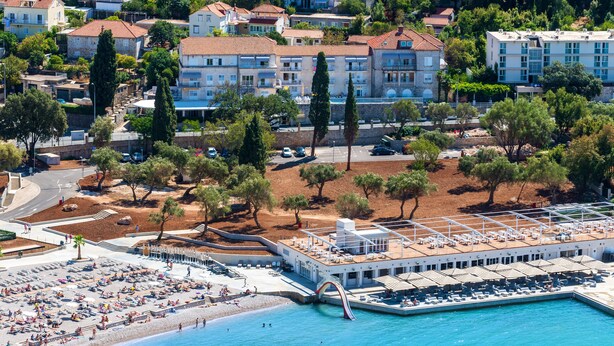 An aerial view of a coastal town with a beach, a pool area, and residential buildings. - Hotel Komodor (Dubrovnik)