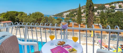 A poolside table with books, croissants, and orange juice glasses, overlooking a pool and a coastal town.