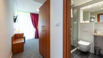 Quarto de hotel com guarda-roupa de madeira, uma mesa de trabalho com abajur, área de chuveiro e casa de banho.