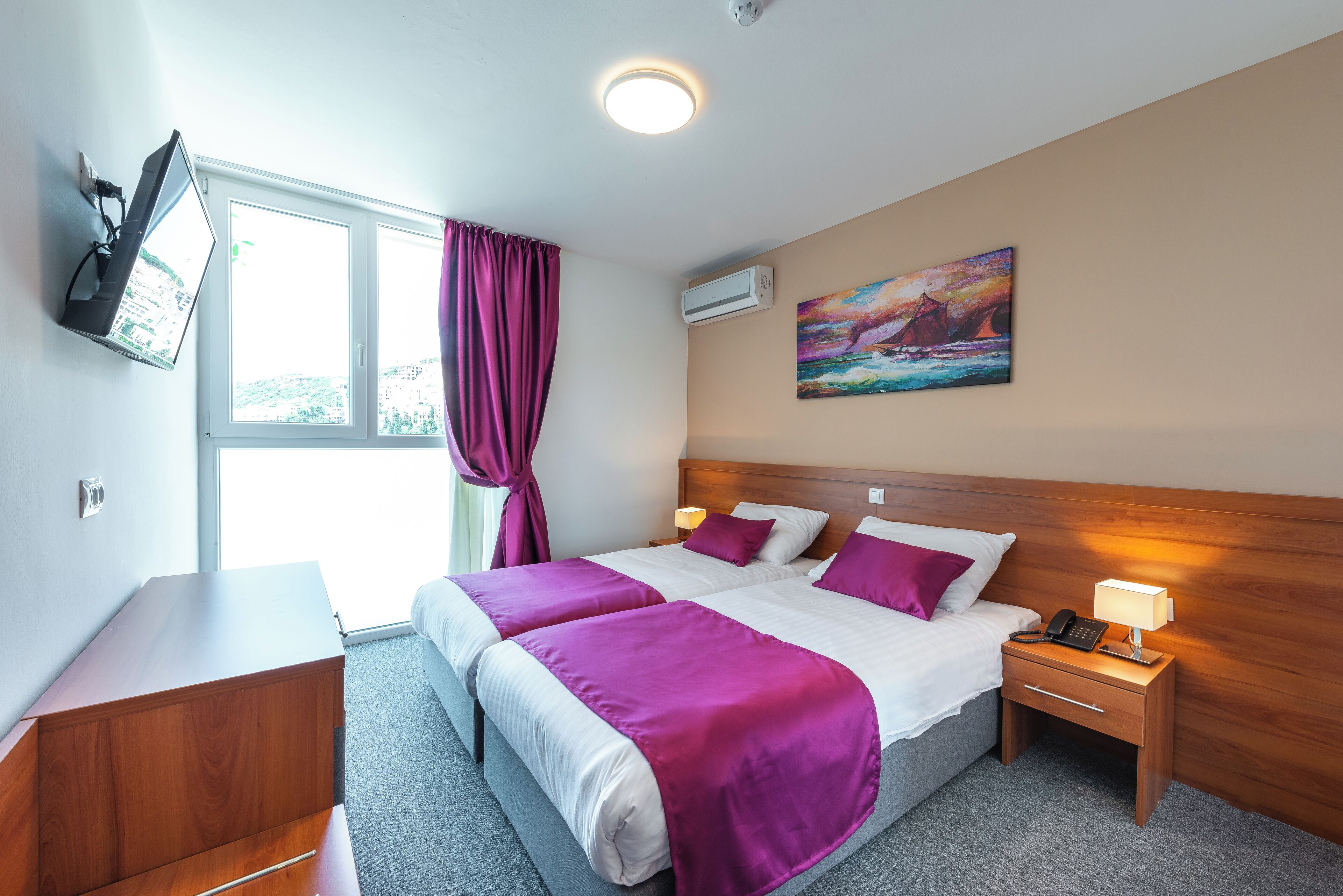 Una habitación de hotel con una cama, una mesita de noche con una lámpara, un televisor montado en la pared y un cuadro en la pared.