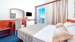 A hotel room with a large bed, a desk, a chair, a TV, and a view of the outdoors. - Hotel Komodor (Dubrovnik)