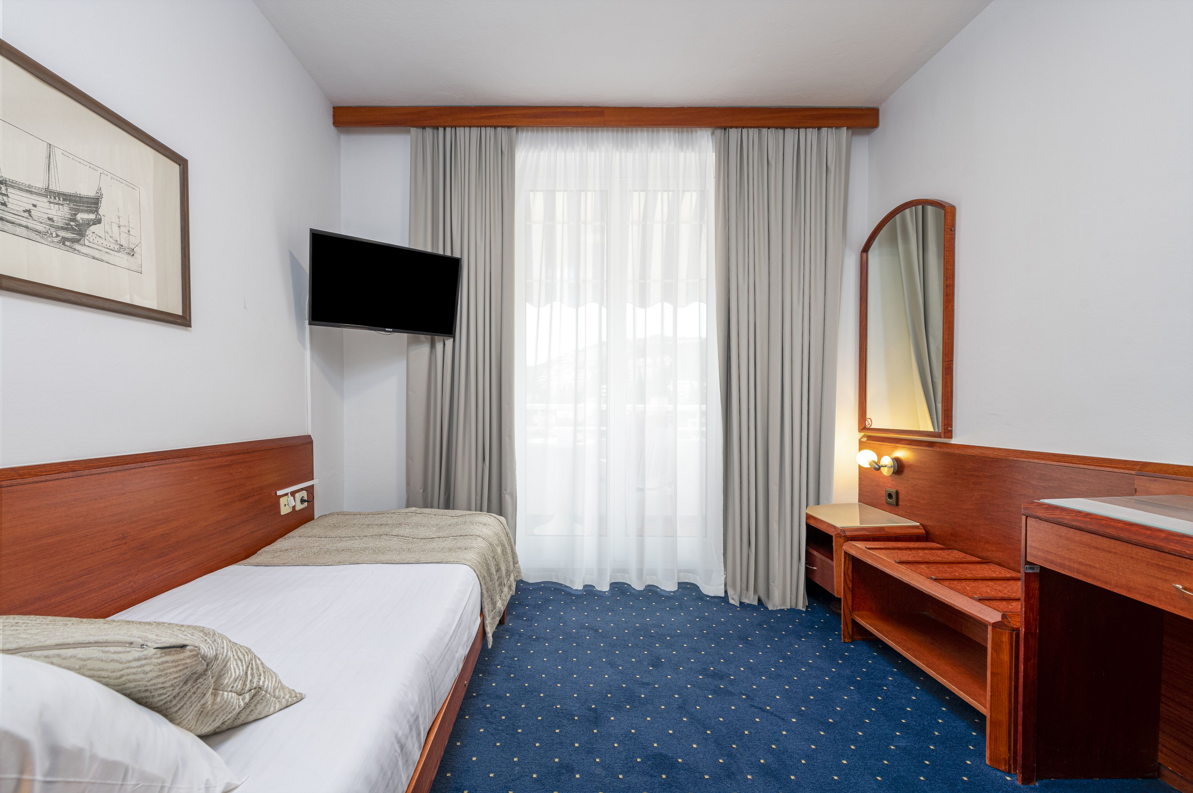 Una habitación de hotel con una cama, televisión, escritorio, espejo y ventana con cortinas.