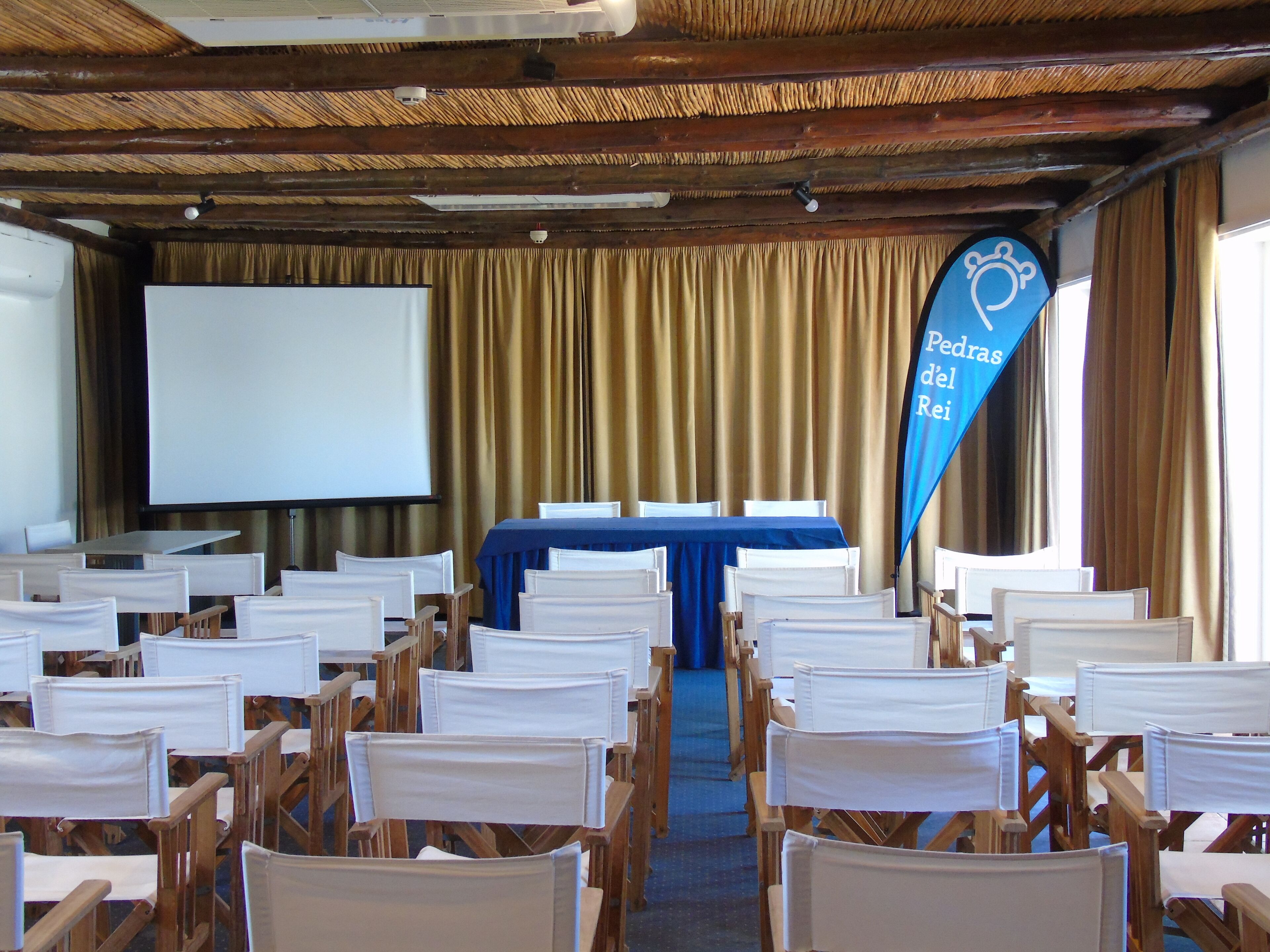 A conference room with rows of wooden chairs, a blue table, a projector screen, and a banner reading "Petras del Rei".