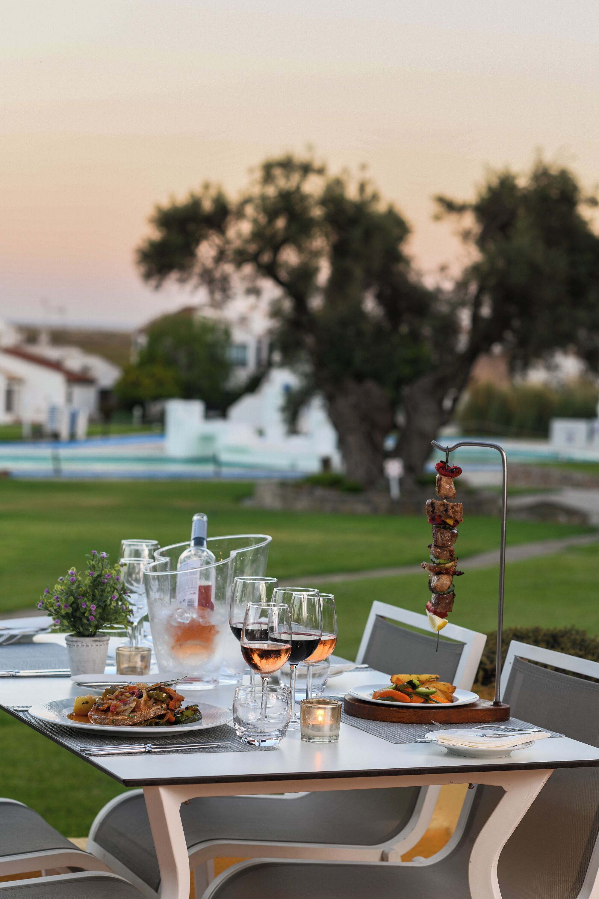 An outdoor dining table set with plates of food, glasses, and a bottle of wine, with a skewer of meat and vegetables in the background.