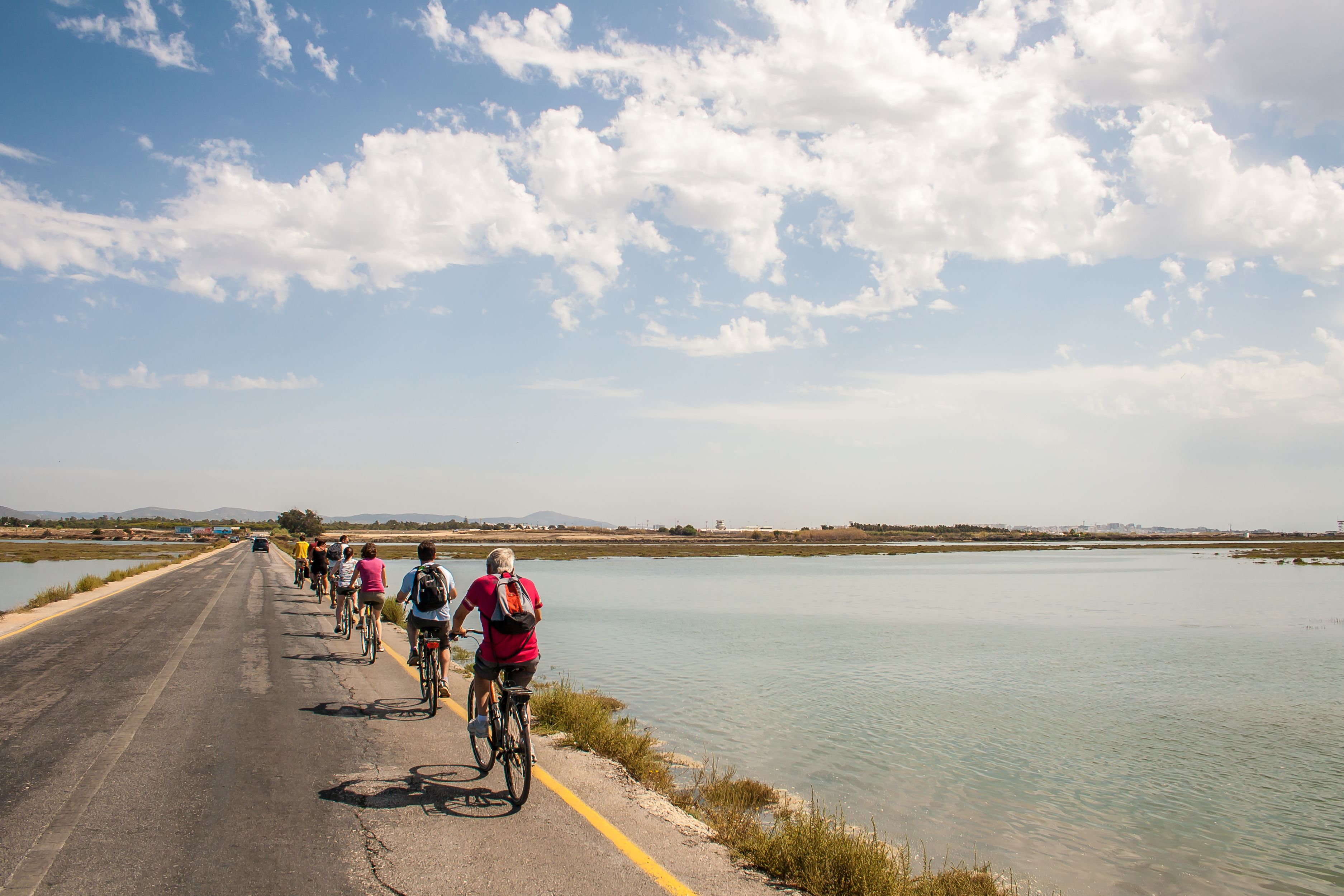 A group of cyclists on a paved road beside a calm body of water, with a clear sky above.