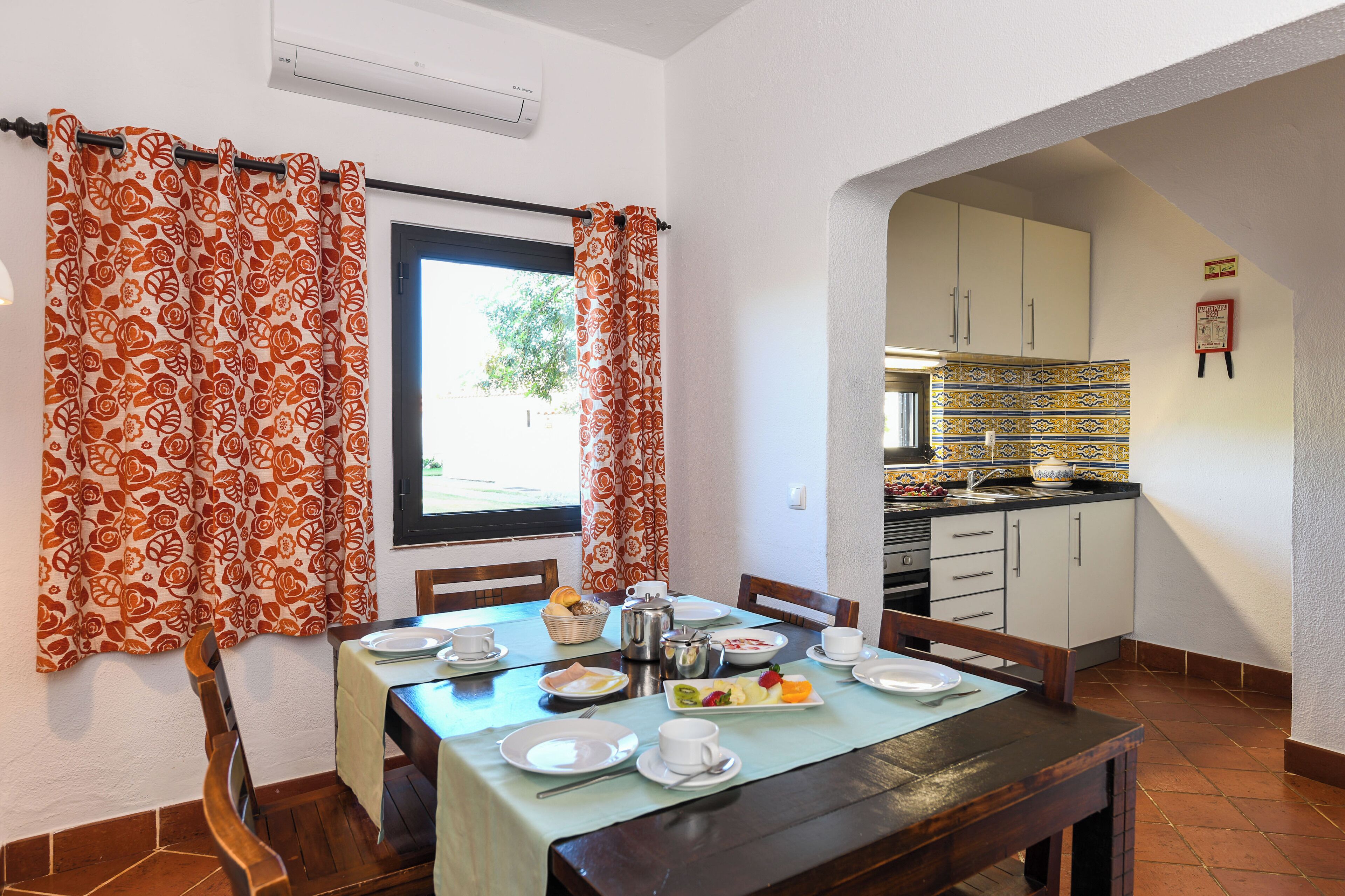 Un comedor con una mesa lista para dos personas, una ventana con cortinas y una cocina al fondo.