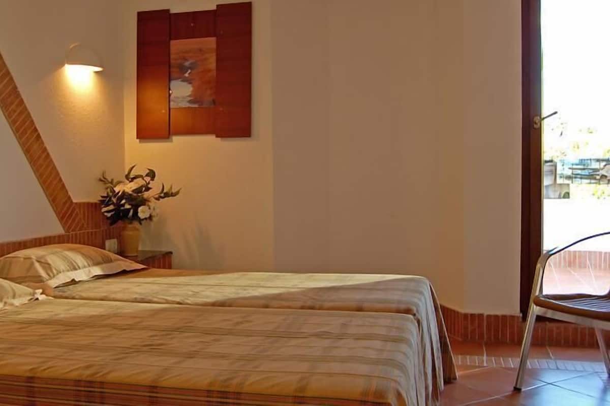 A room with two beds, a wall-mounted light, a painting, and a potted plant.