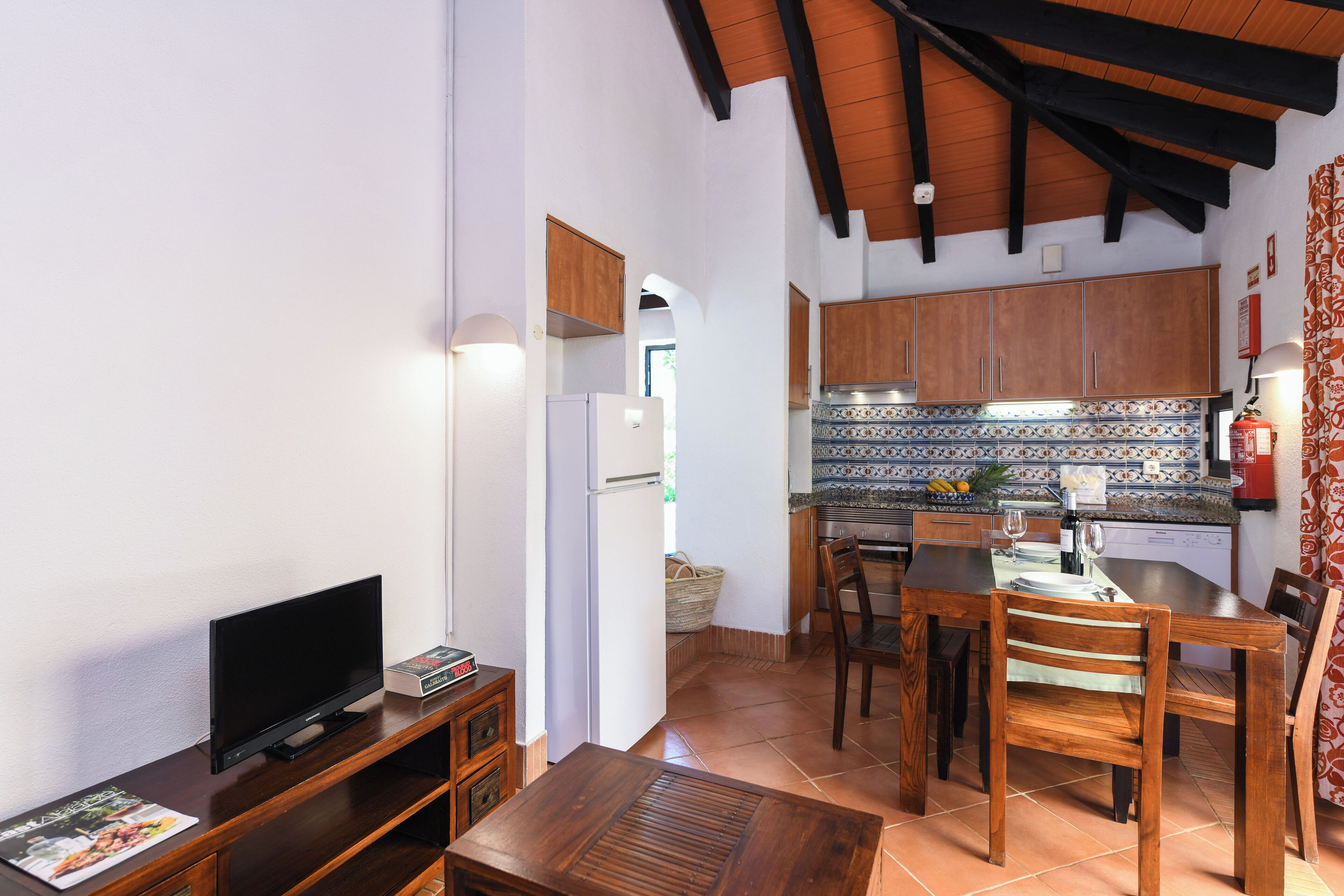 A compact living space with a television on a wooden stand, a dining table with chairs, and a kitchen area with cabinets and a tiled backsplash.