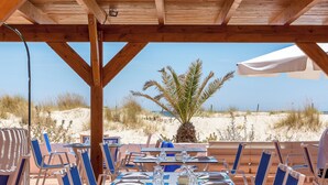 An outdoor dining area with tables and chairs, a palm tree, and a view of the beach and dunes. - Pedras d'el Rei (Tavira)