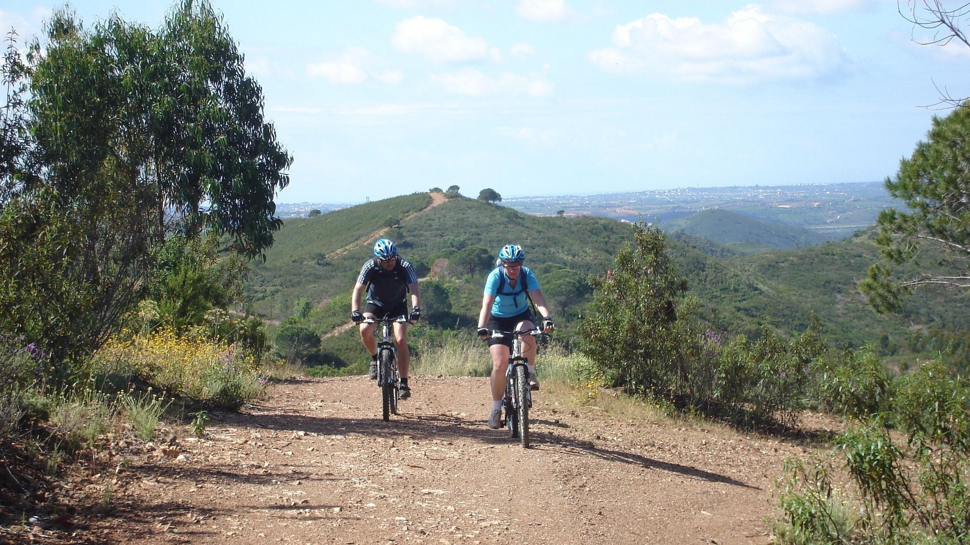 Two cyclists on a dirt path surrounded by greenery and hills.