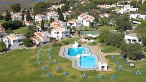 An aerial view of a residential area with white houses, a large swimming pool, and numerous blue lounge chairs. - Pedras d'el Rei (Tavira)