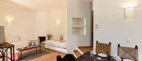 Een moderne woonkamer met een bank, een salontafel en een televisie. Er is een eethoek met een tafel gedekt voor twee personen, stoelen en een open haard.