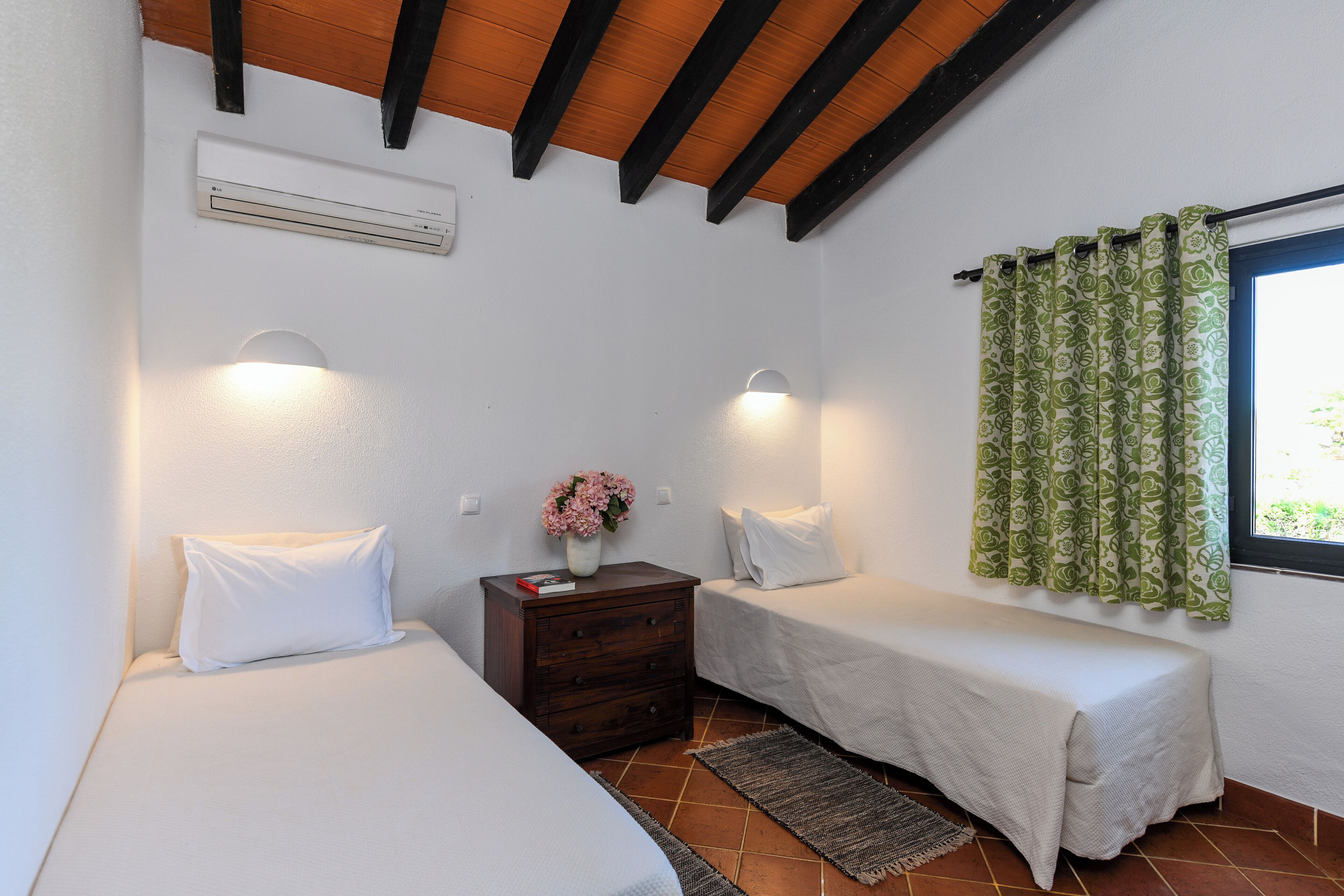 A bedroom with two single beds, a nightstand with a vase of flowers, and a window with patterned curtains.