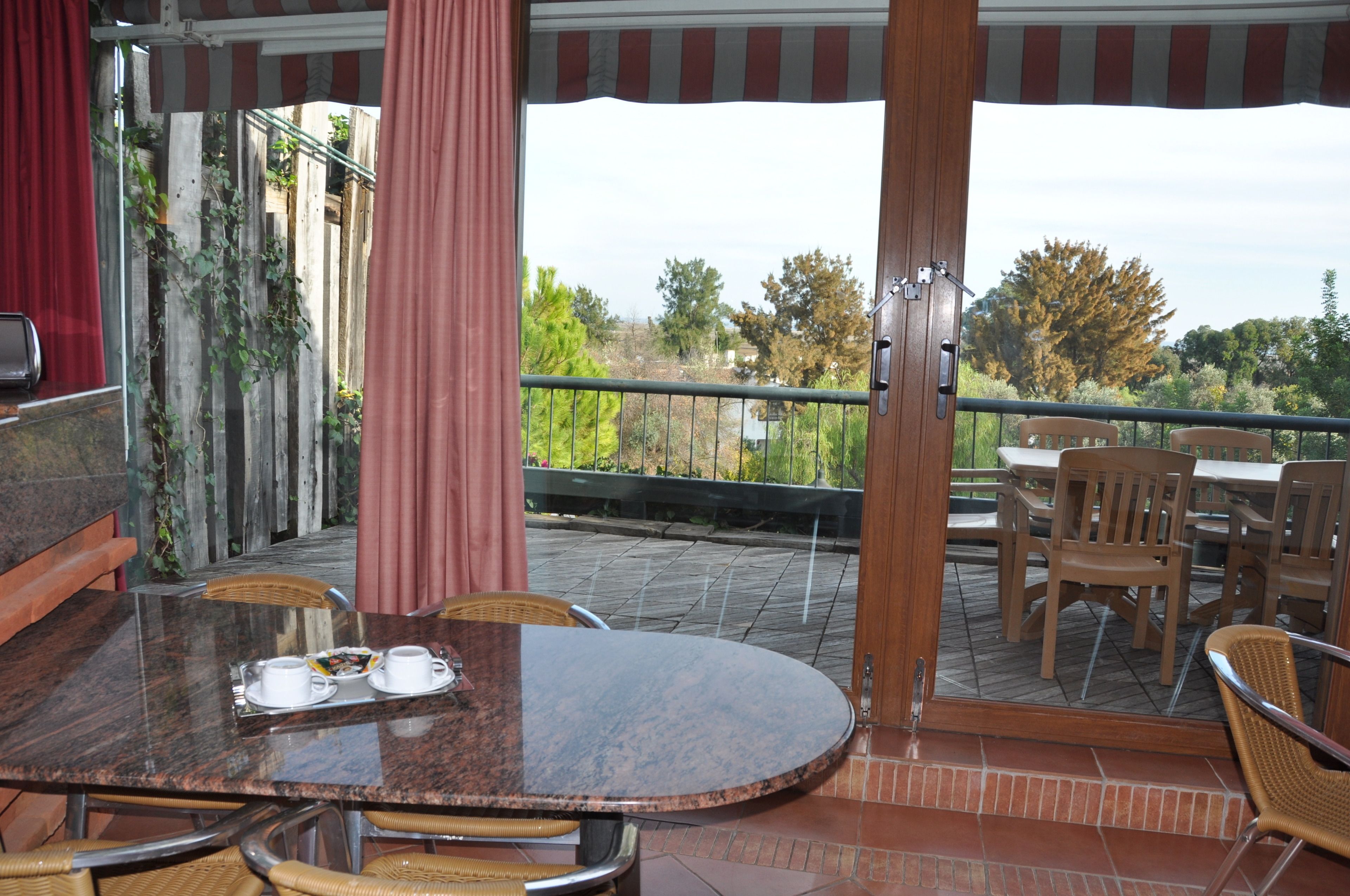A covered outdoor dining area with a table and chairs, a view of the surrounding landscape, and a wooden fence with climbing plants.