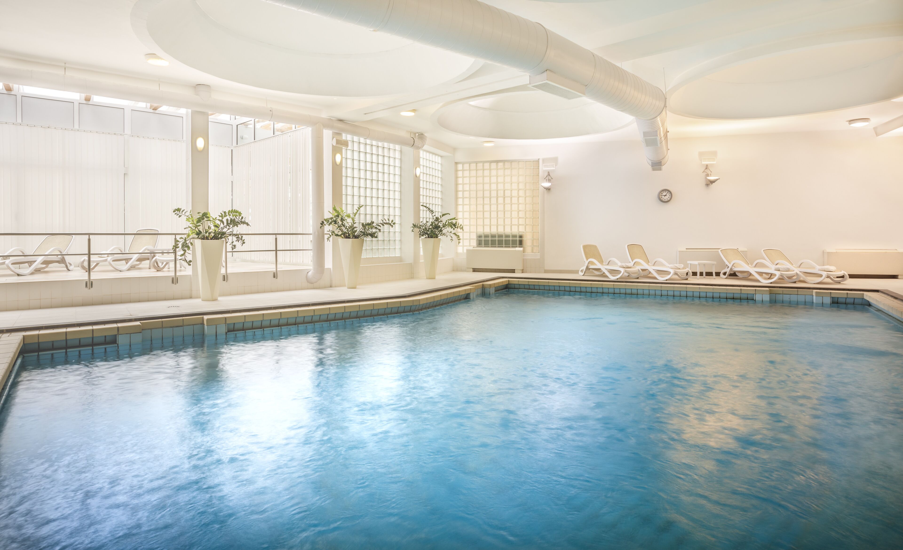 Une piscine intérieure à l’eau d’un bleu cristallin, avec des chaises longues blanches et des plantes en pot.