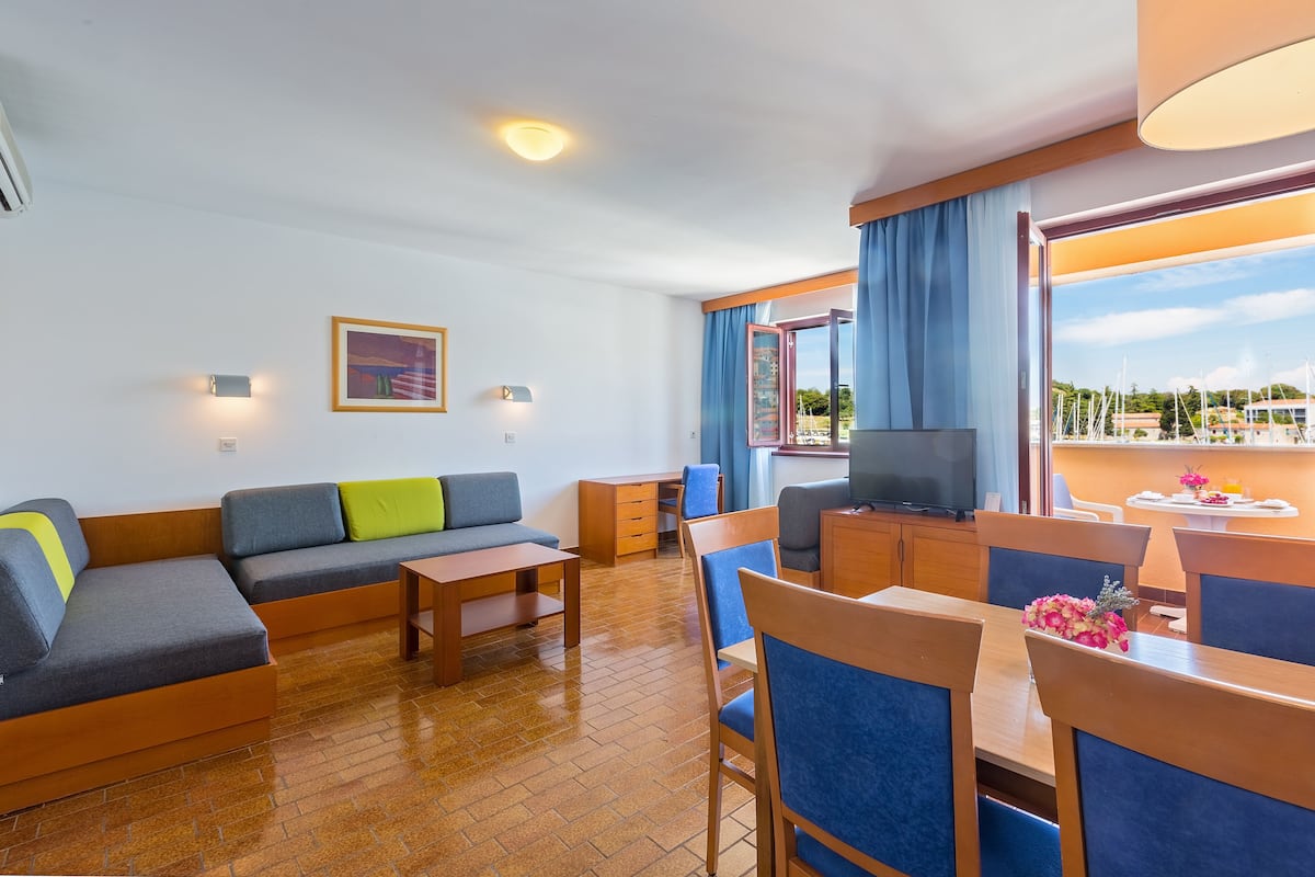 A modern hotel room with a sofa, a desk, a TV, and a dining area with chairs and a table.