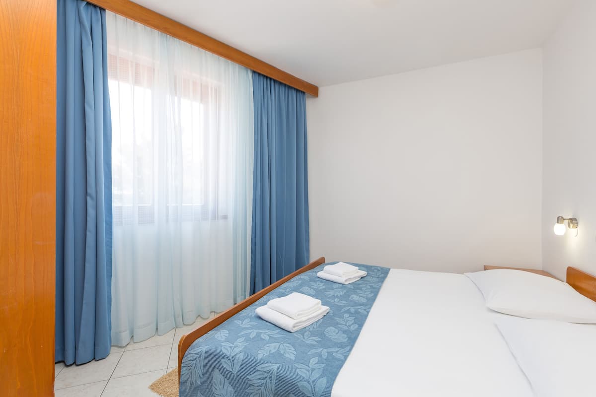 A hotel room with a bed, a wooden headboard, a window with blue curtains, and a wall-mounted light fixture.