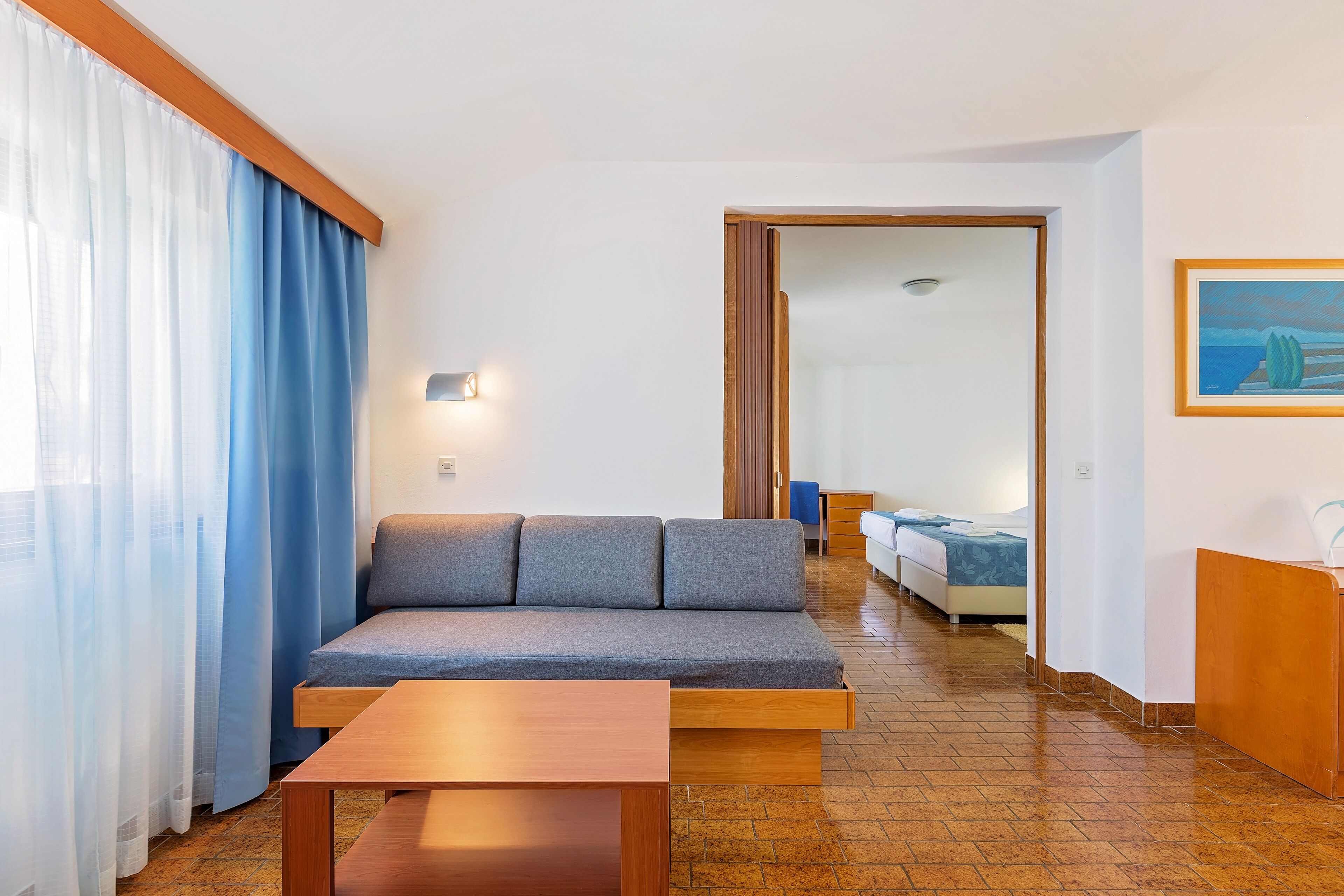 A modern hotel room with a sofa, a wooden table, a bed, and a painting on the wall.