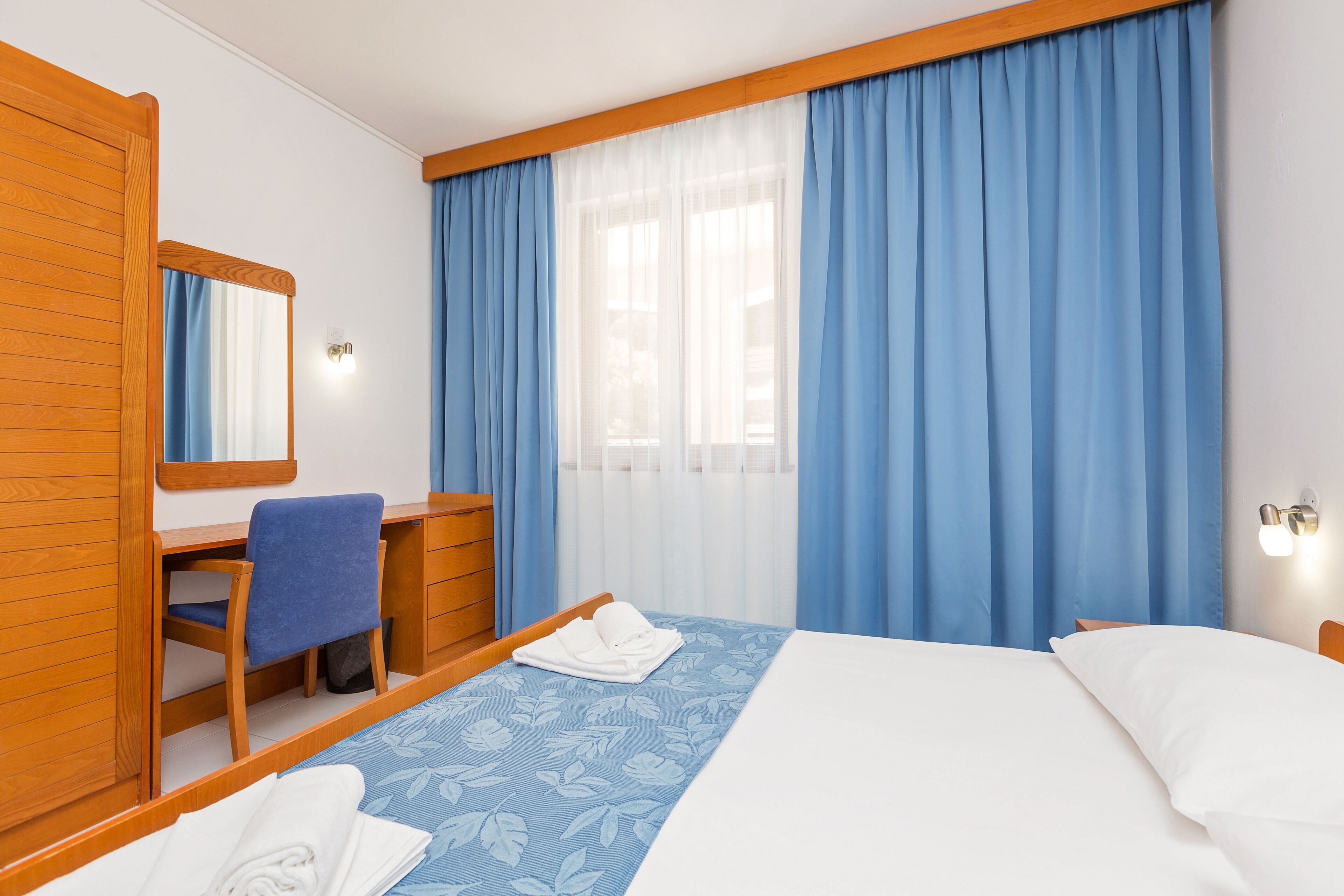 A hotel room with a bed, a desk, a chair, and a large window with blue curtains.