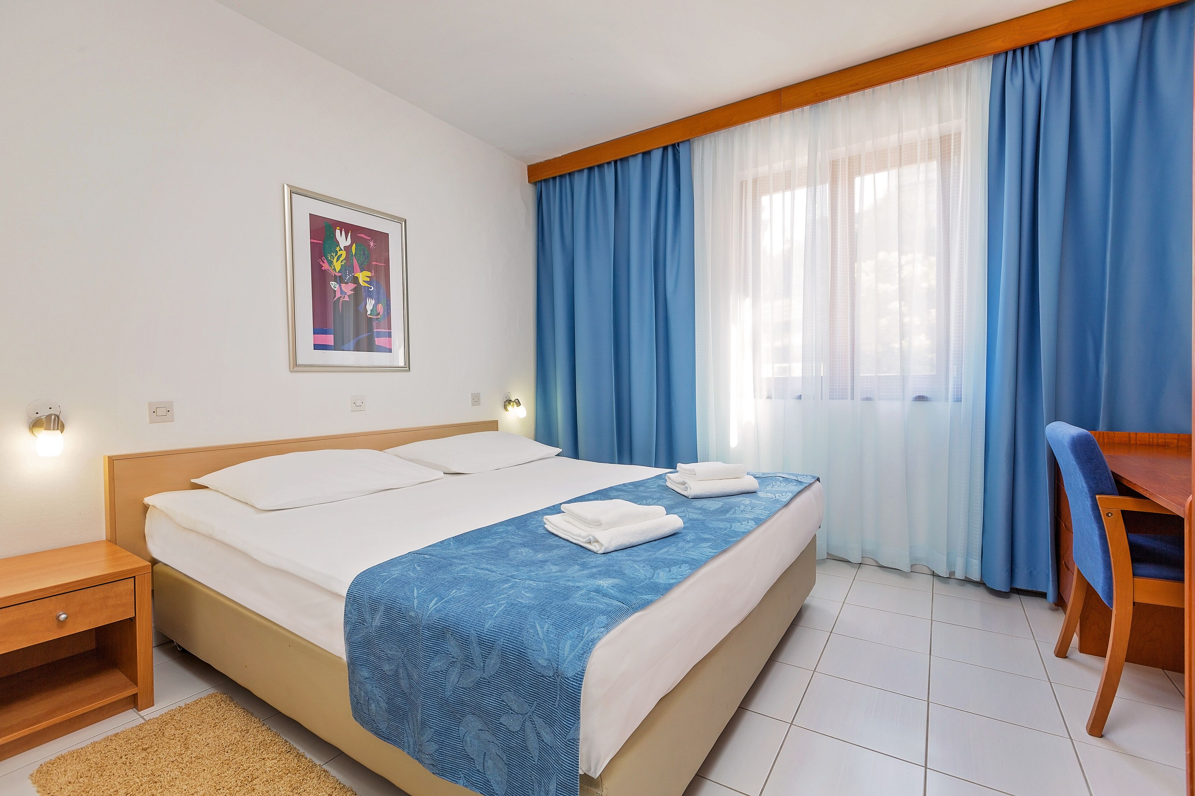 A hotel room with a bed, a desk, a chair, a framed picture on the wall, and a window with blue curtains.