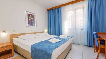 A hotel room with a bed, a desk, a chair, a framed picture on the wall, and a window with blue curtains.
