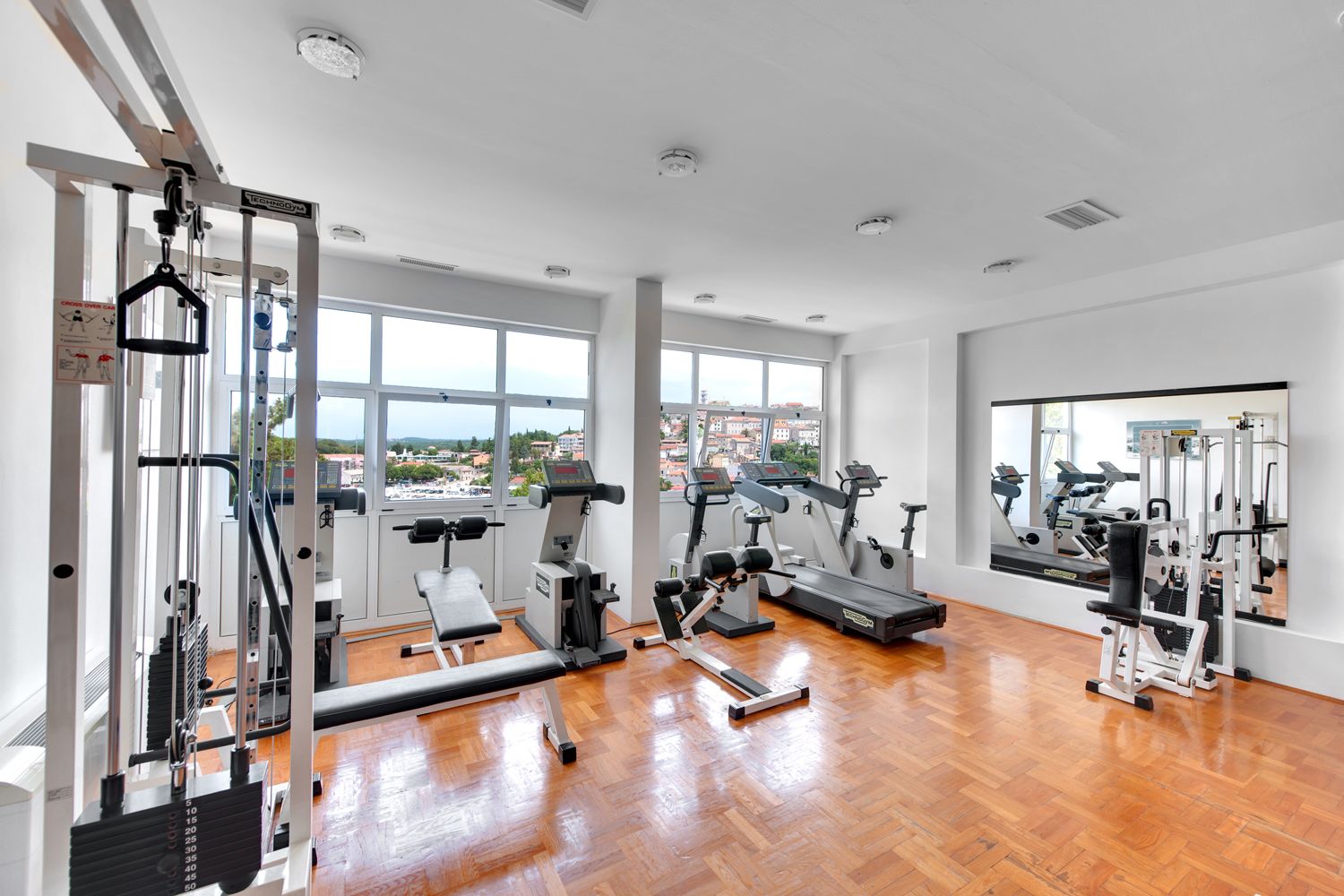 Een ruime fitnessruimte met diverse apparaten, grote ramen en uitzicht op gebouwen.