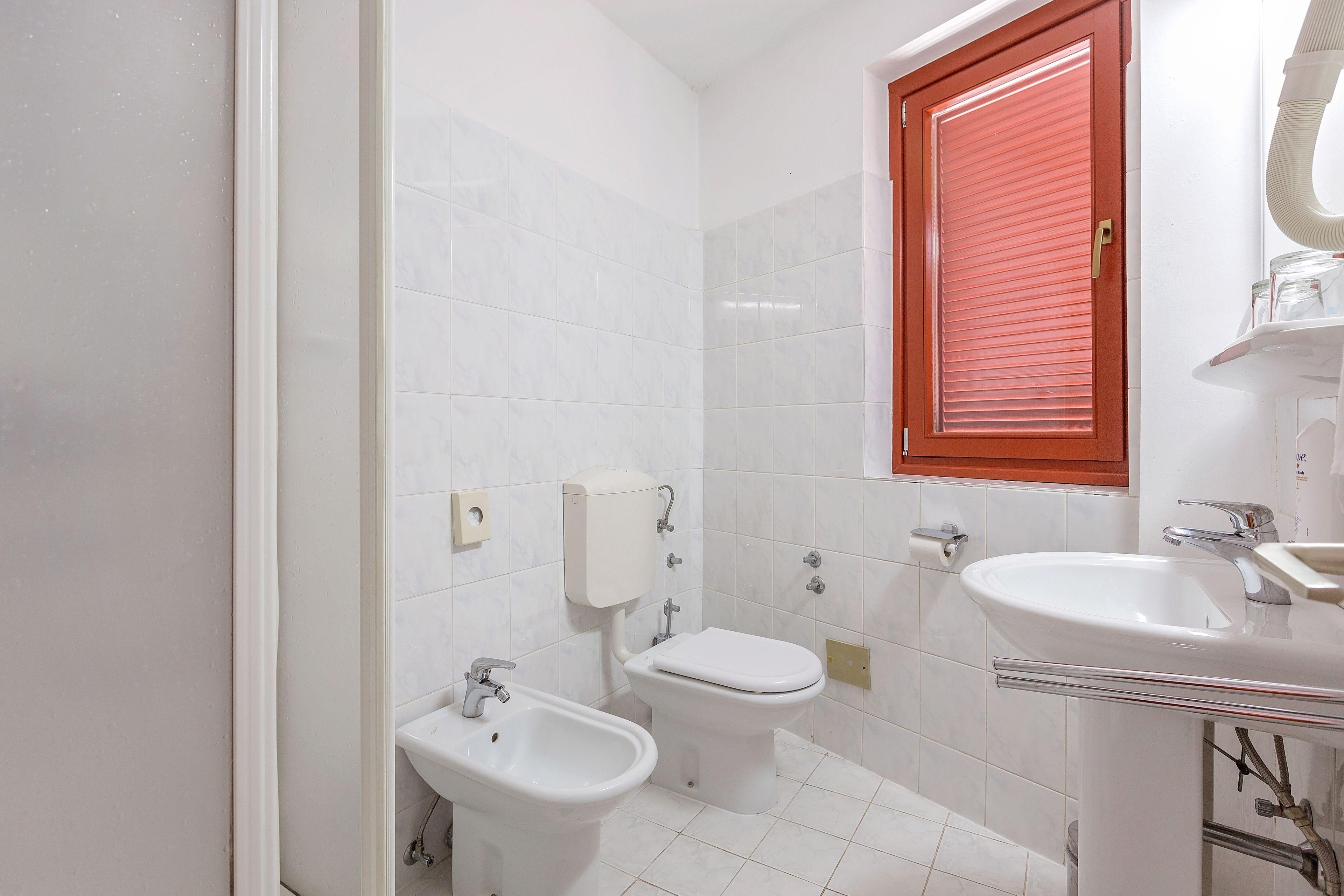 Ein Badezimmer mit weißen Fliesen, einem roten Fensterladen, einem Bidet, einer Toilette und einem Waschbecken.