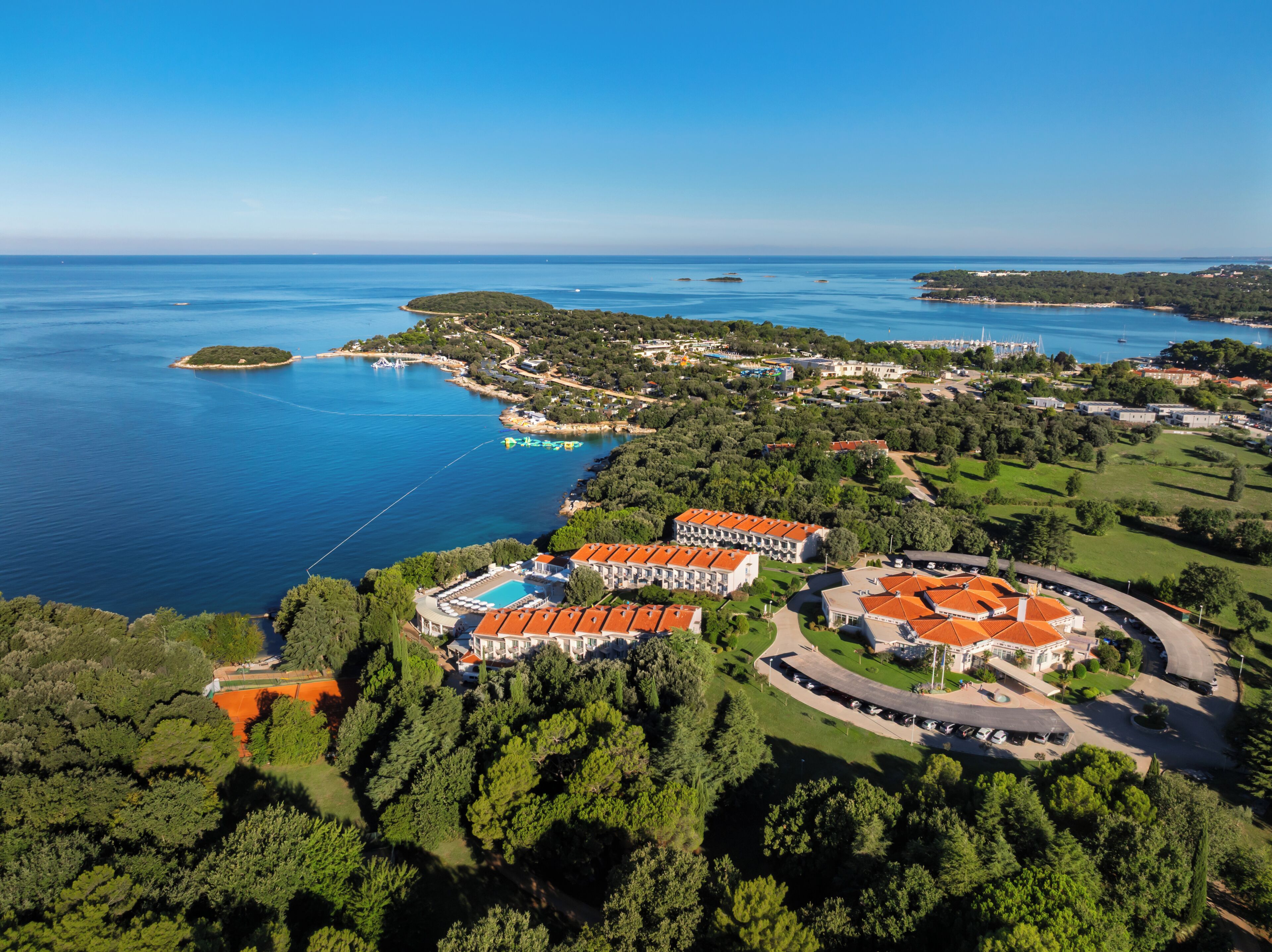 An aerial view of a coastal resort with multiple buildings, a swimming pool, and surrounding greenery.