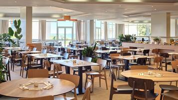 A spacious dining area with multiple tables and chairs, large windows with curtains, and potted plants.