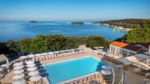 A poolside area with lounge chairs and umbrellas, a clear view of the sea, and a building with a red roof. - Maistra Select Funtana All Inclusive Resort (Funtana)