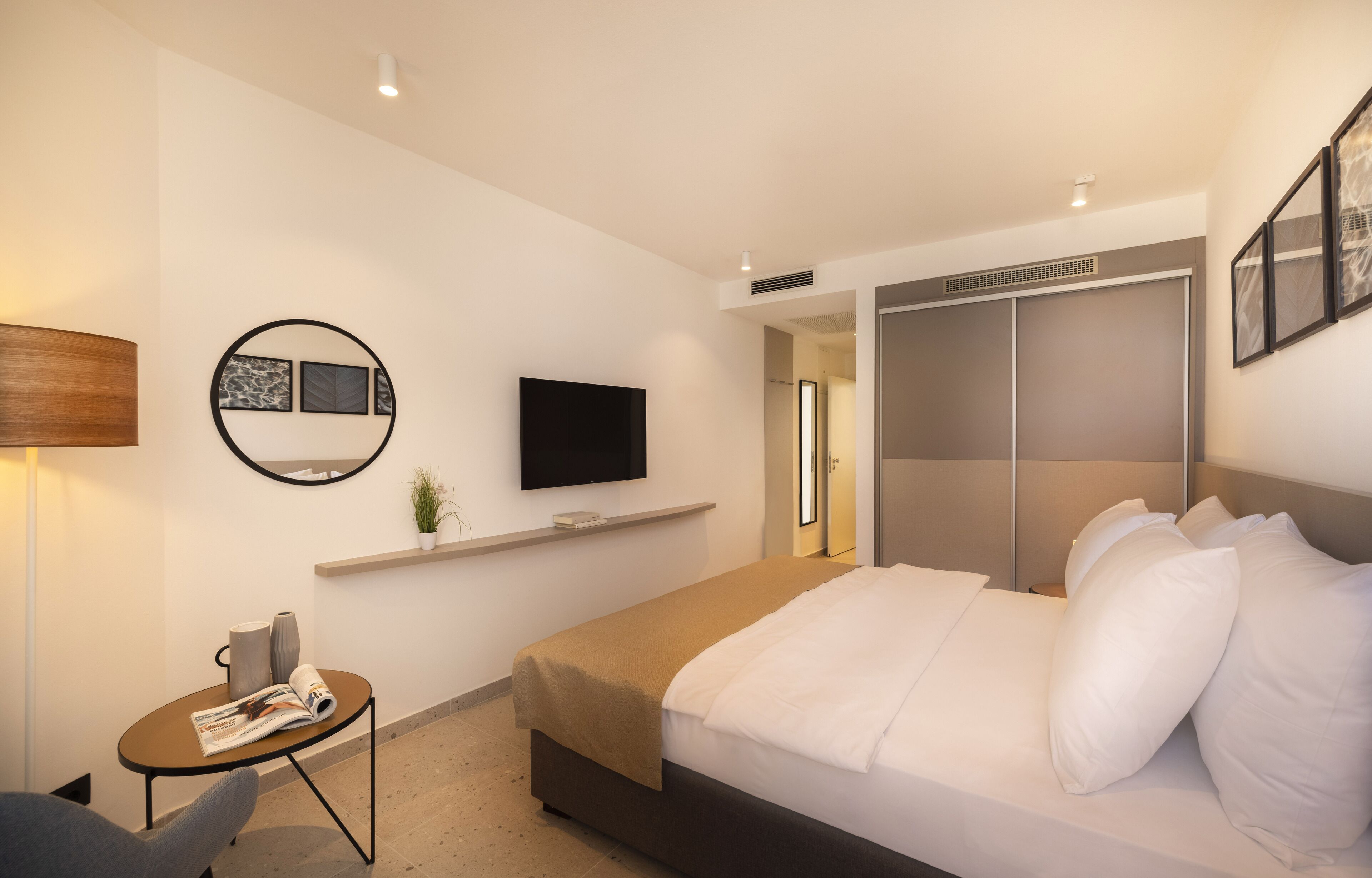 A modern hotel room with a bed, a TV mounted on the wall, a small table with a magazine, and a closet with sliding doors.