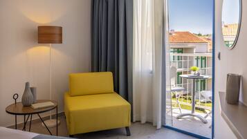 Habitación de hotel con un sillón amarillo, una mesita redonda con lámpara y vistas a un balcón con mobiliario exterior.