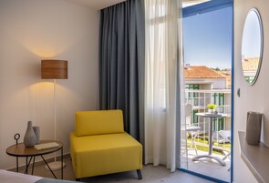 Habitación de hotel con un sillón amarillo, una mesita redonda con lámpara y vistas a un balcón con mobiliario exterior.