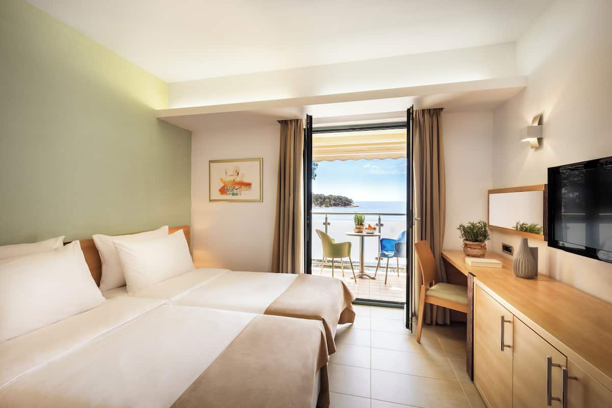 Une chambre d’hôtel avec deux lits, un bureau, une télévision et un balcon donnant sur la mer.