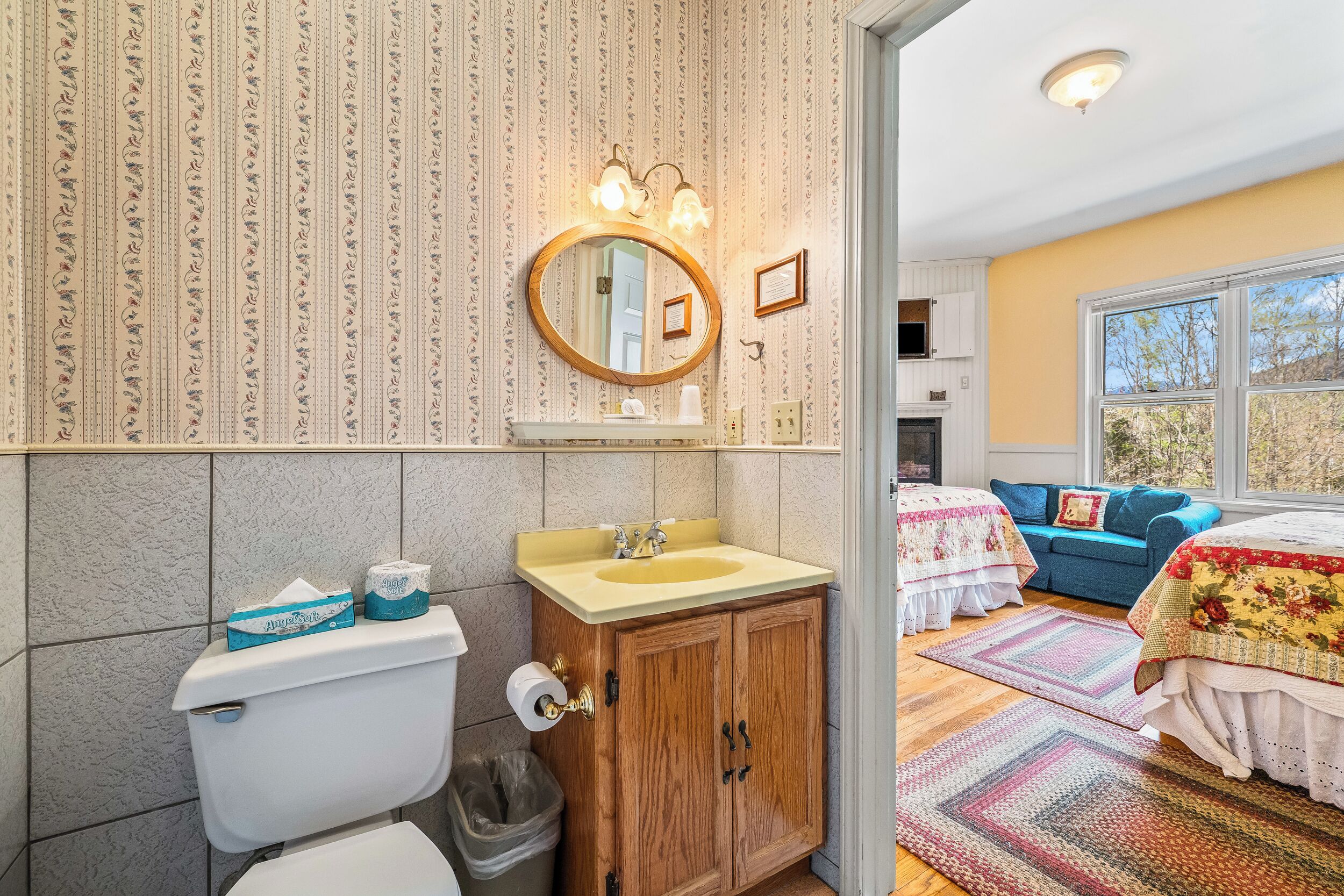 A bathroom with a toilet, sink, and mirror, wooden cabinets, and a window with a view.