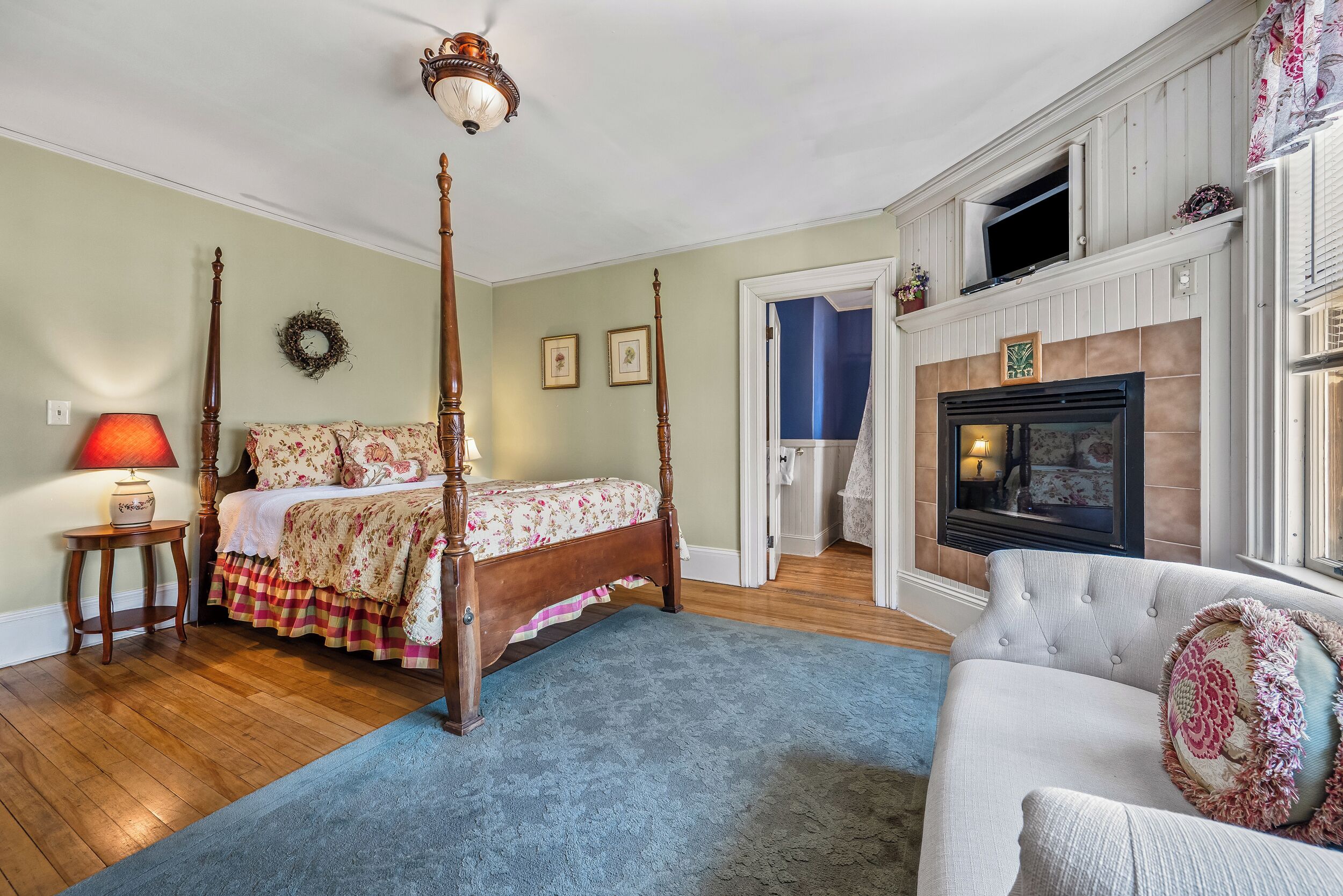 A bedroom with a four-poster bed, a fireplace, a blue carpet, and a white armchair.