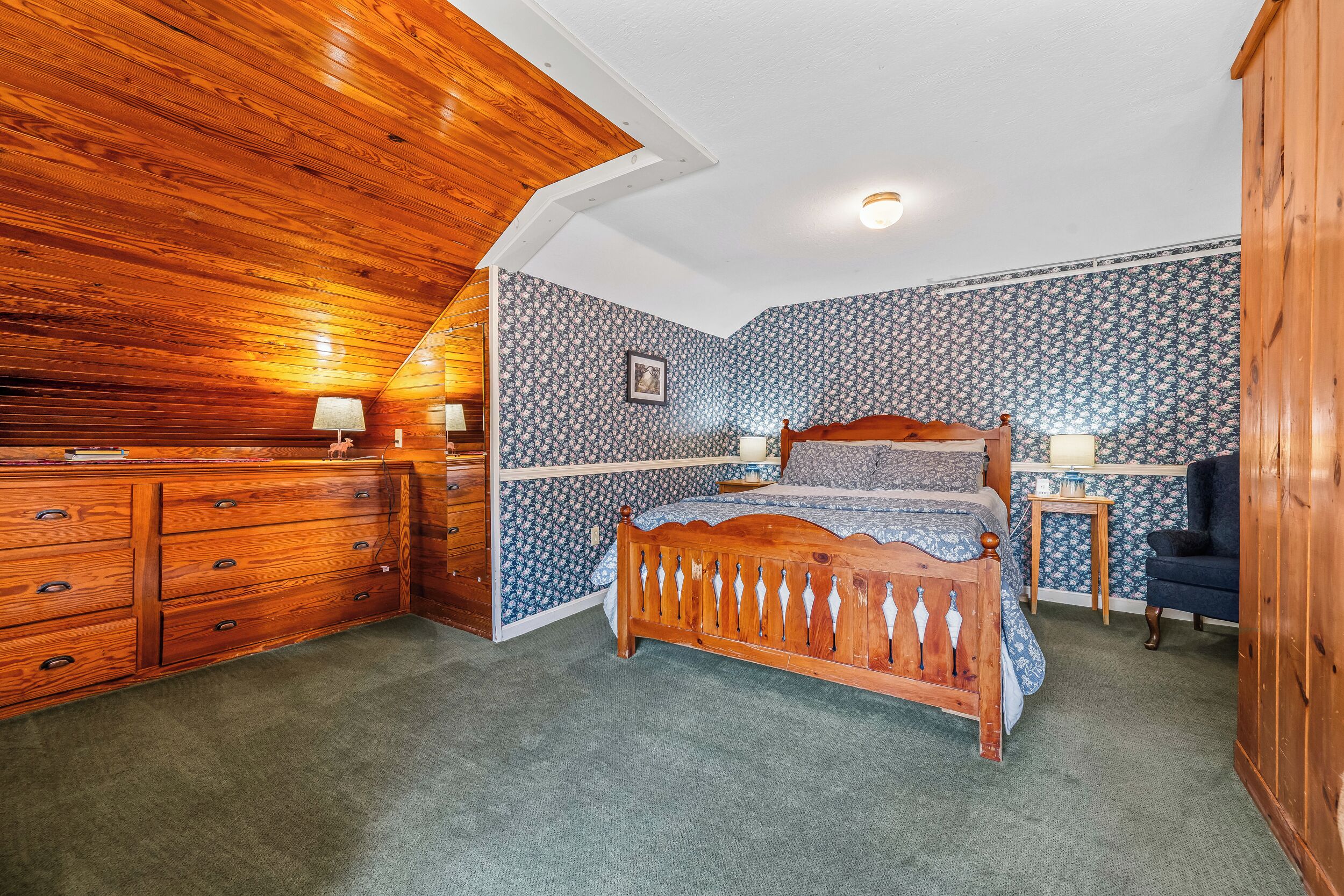 A bedroom with a wooden ceiling, a bed with a wooden headboard, a dresser, and patterned wallpaper.