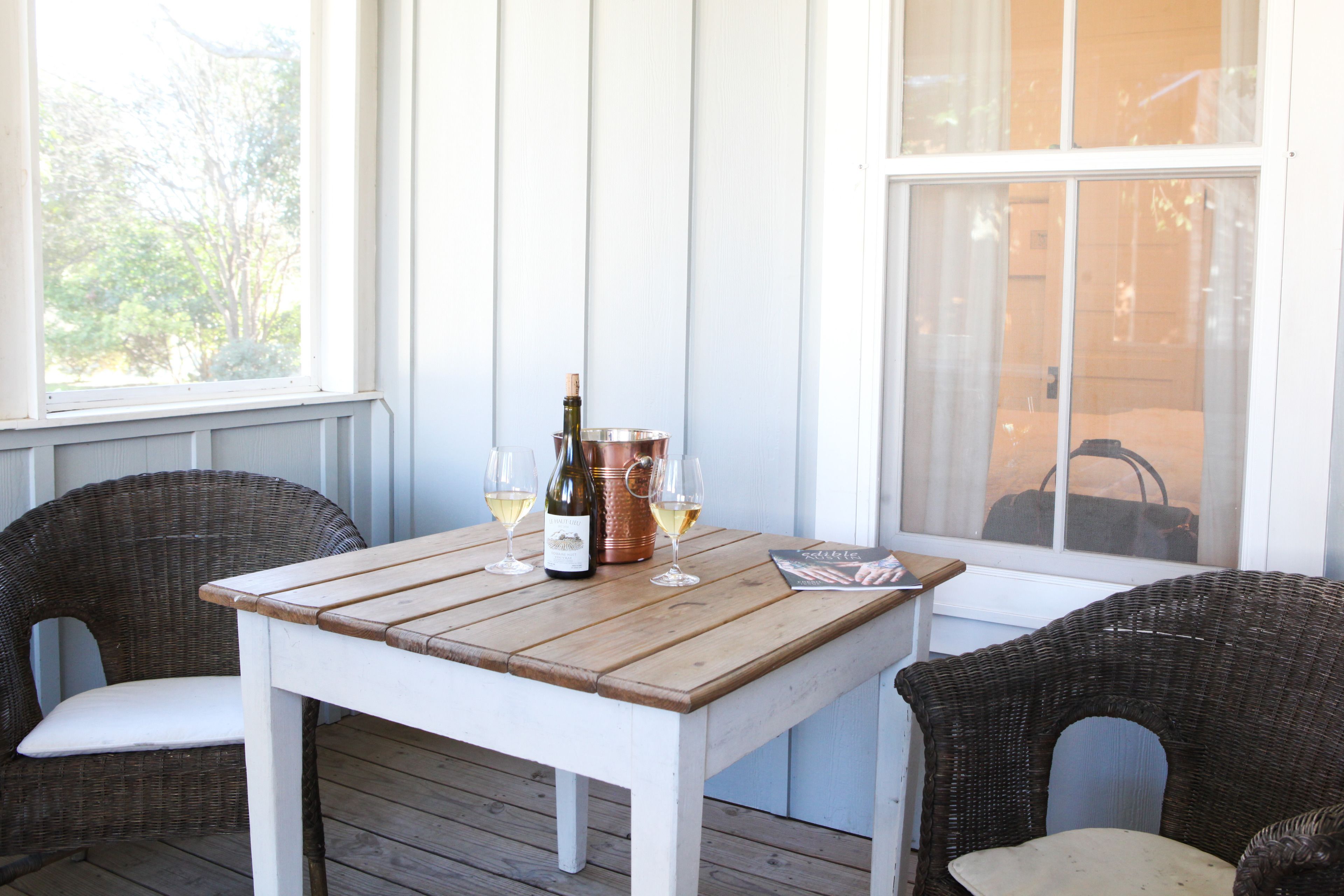 A wooden table with a bottle of wine and glasses, surrounded by wicker chairs.