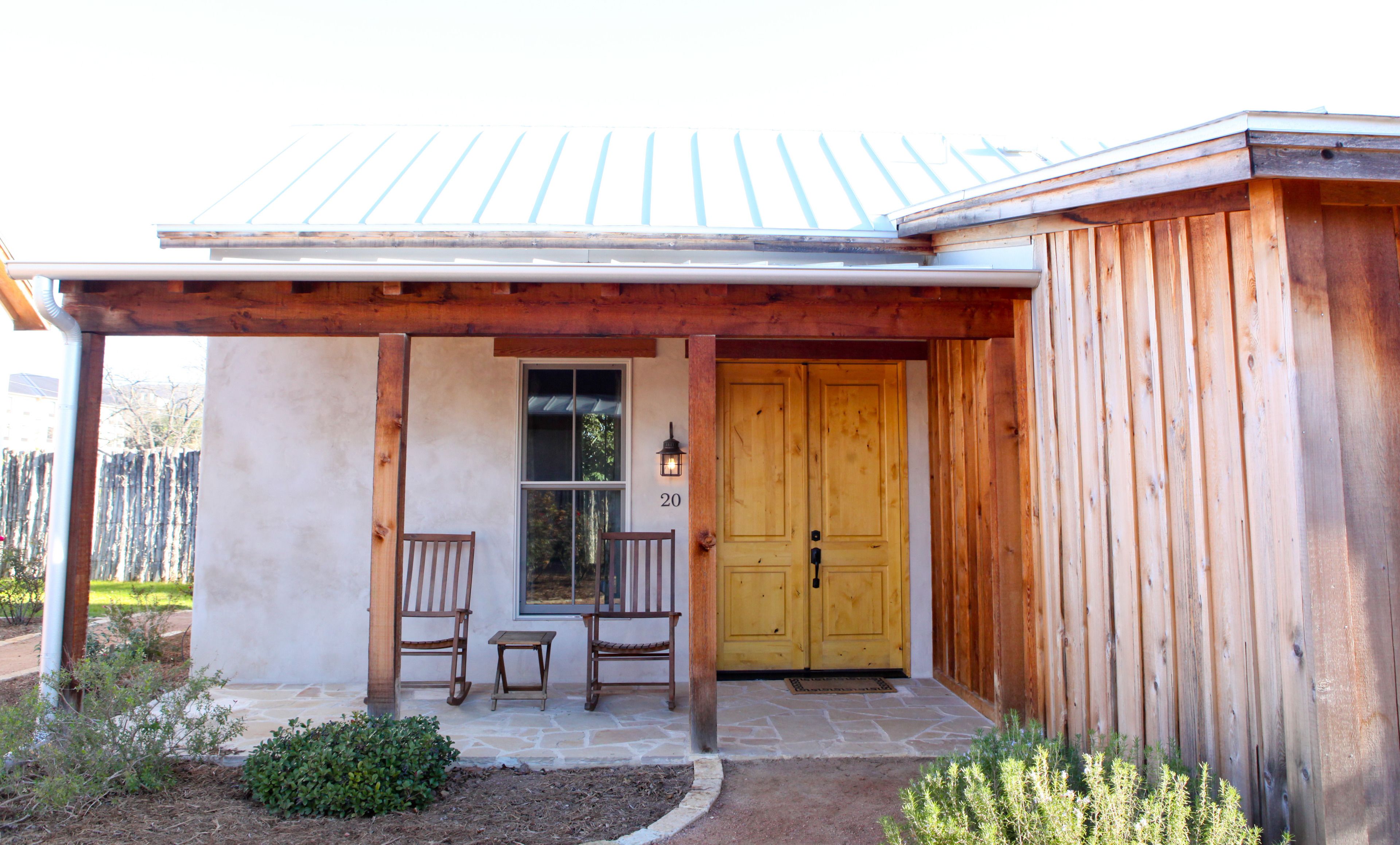 A wooden house with a covered porch, two wooden chairs, a small table, and a yellow door.