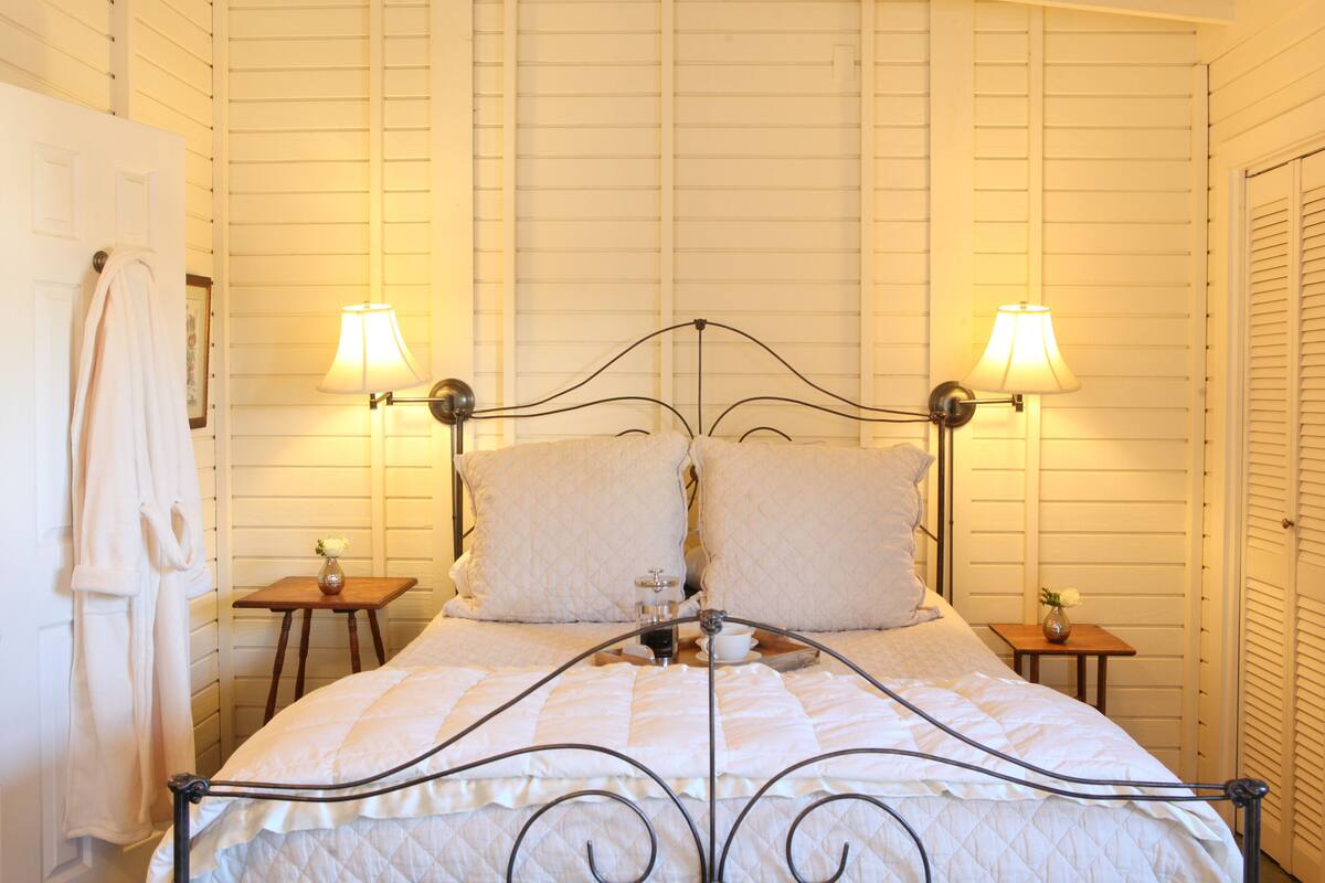 A neatly made bed with a metal frame and white bedding, two bedside tables with lamps, and a white robe hanging on a door.