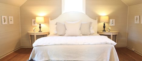 Un dormitorio con cama blanca, dos mesitas de noche con lámparas, una ventana y suelo de madera.