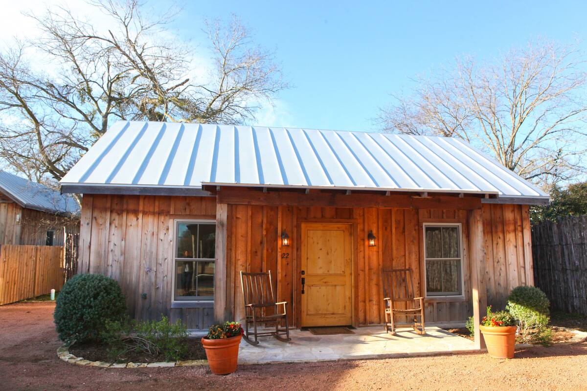 A wooden cabin with a metal roof, a front door, two windows, and a porch with chairs.