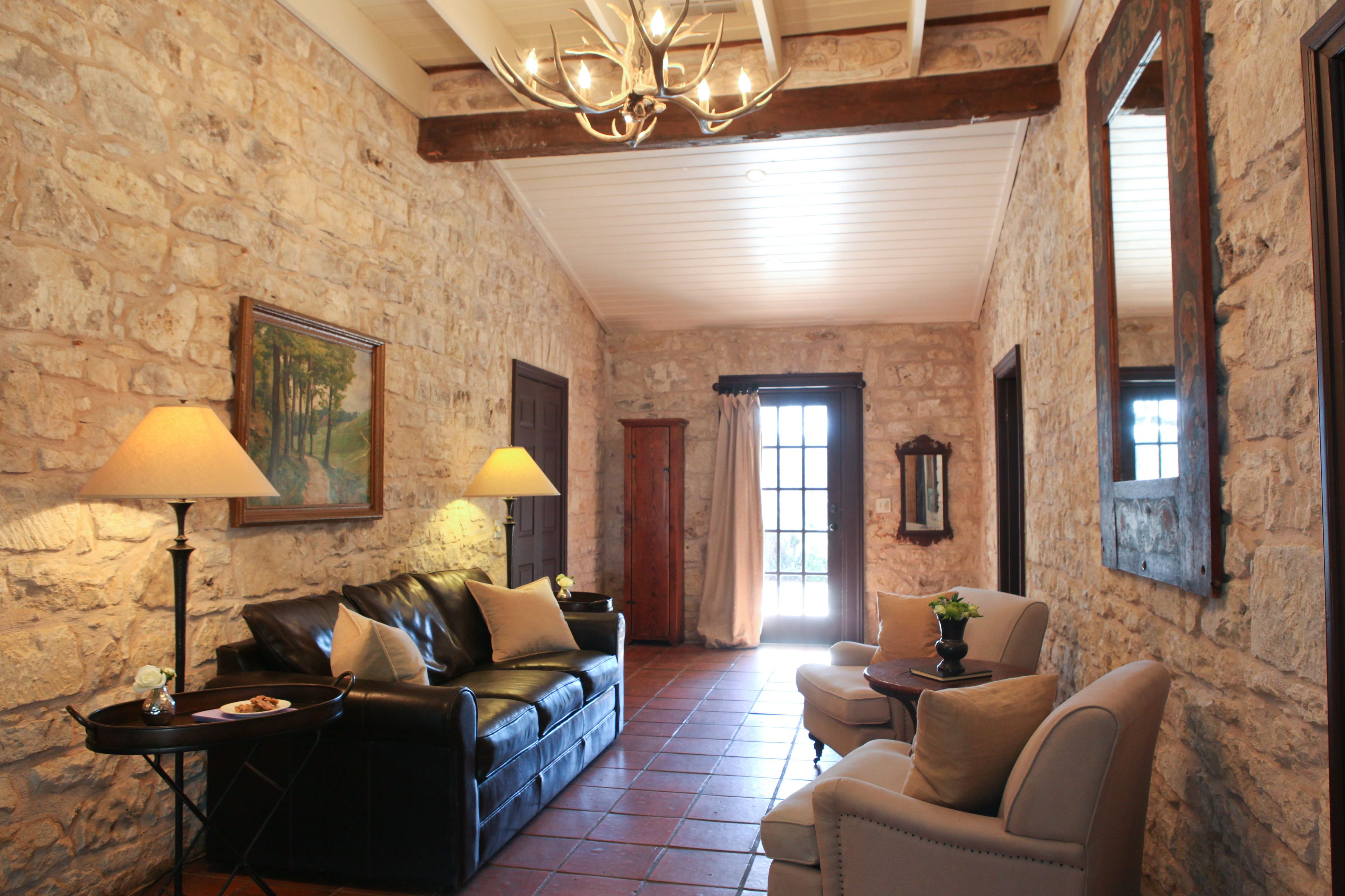 A stone-walled room with a leather sofa, two armchairs, a small table with a lamp, and a chandelier.
