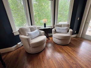 A room with two beige armchairs, a small table with a lamp, and large windows with a view of trees.