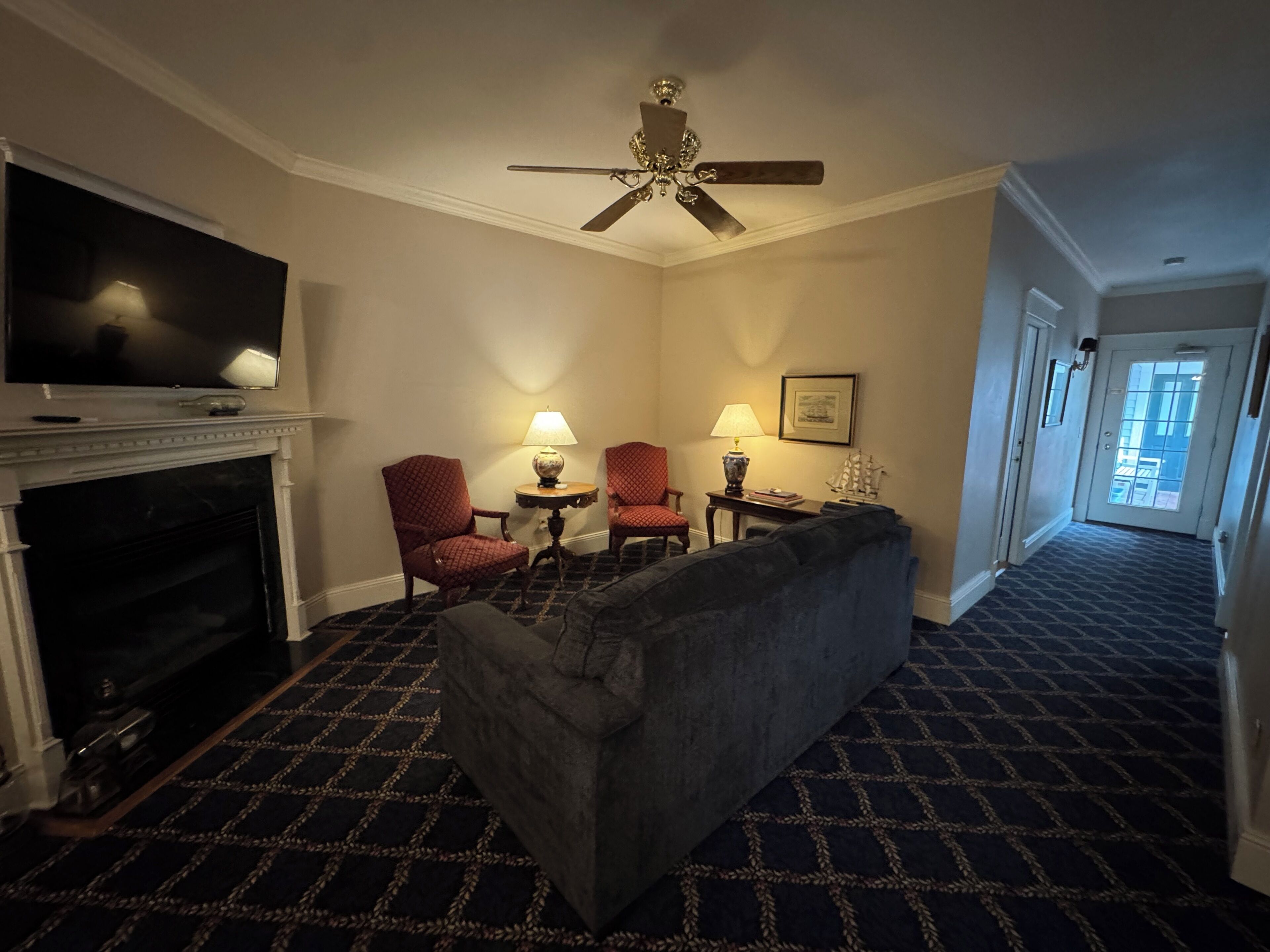 Une chambre d’hôtel équipée d’une cheminée, d’une télévision, d’un ventilateur de plafond, d’un canapé, de deux fauteuils, d’une table d’appoint avec des lampes et d’un tableau encadré au mur.