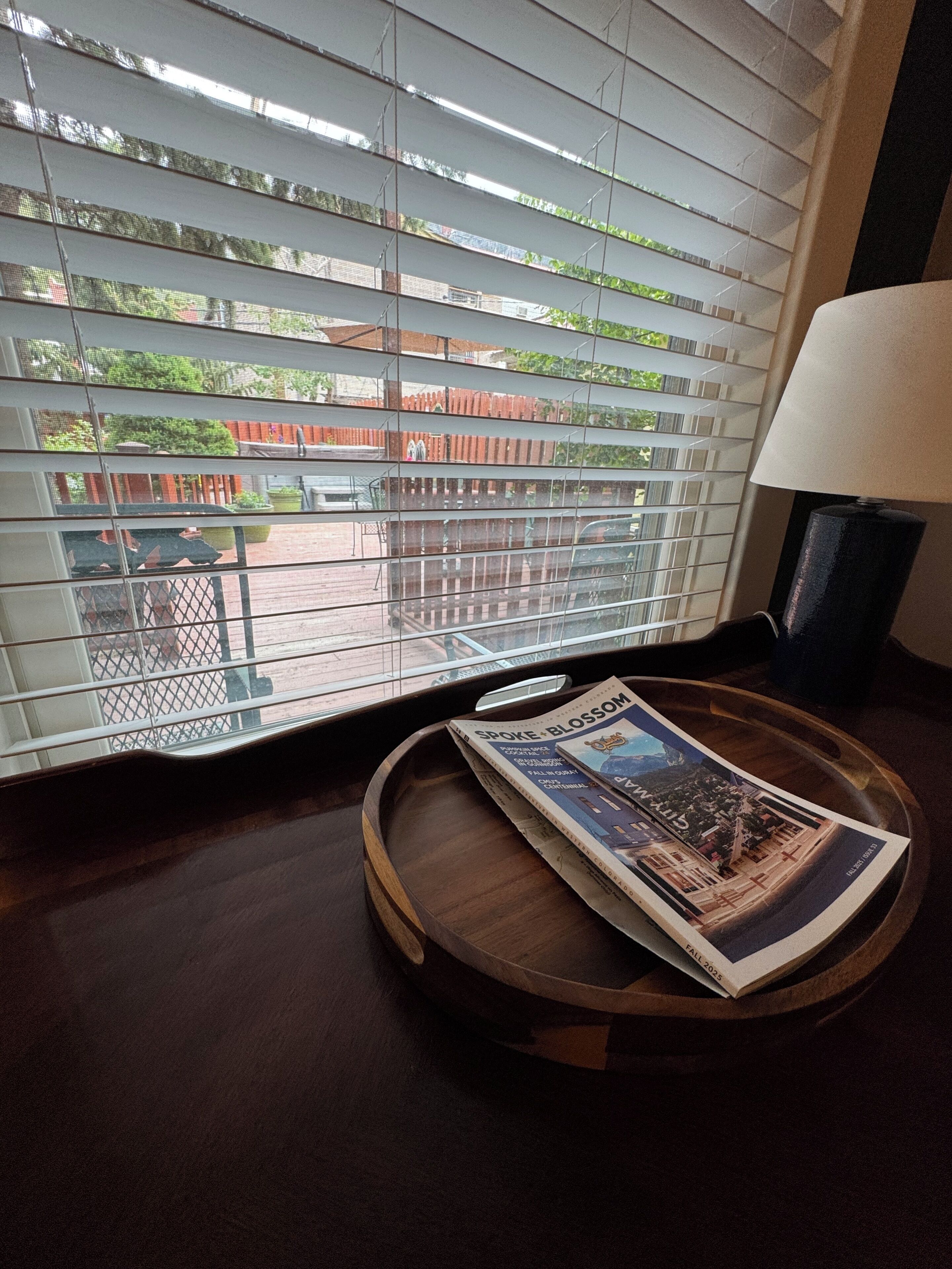 A window with horizontal blinds, a wooden tray with a magazine, and a lamp on a table.