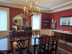 A dining room with a large table, ornate chairs, a chandelier, a mirror, and a wall-mounted shelf with glasses.